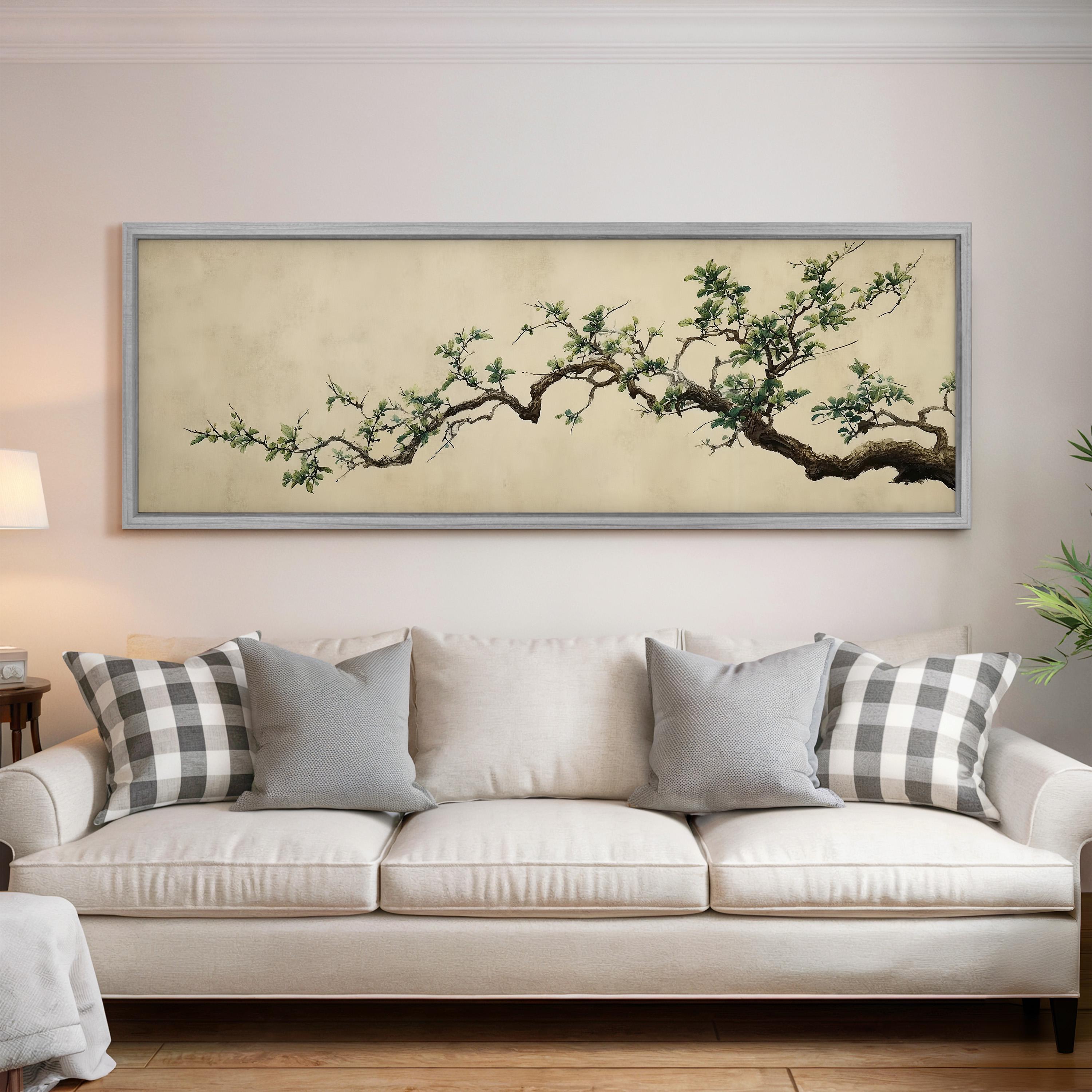 a large, framed painting of a tree branch with green leaves hanging above a white couch with gray and white plaid throw pillows.