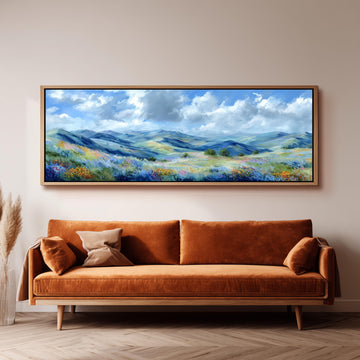 a large, framed painting of a mountainous landscape with a blue sky and fluffy clouds, hanging on a wall above a brown sofa.