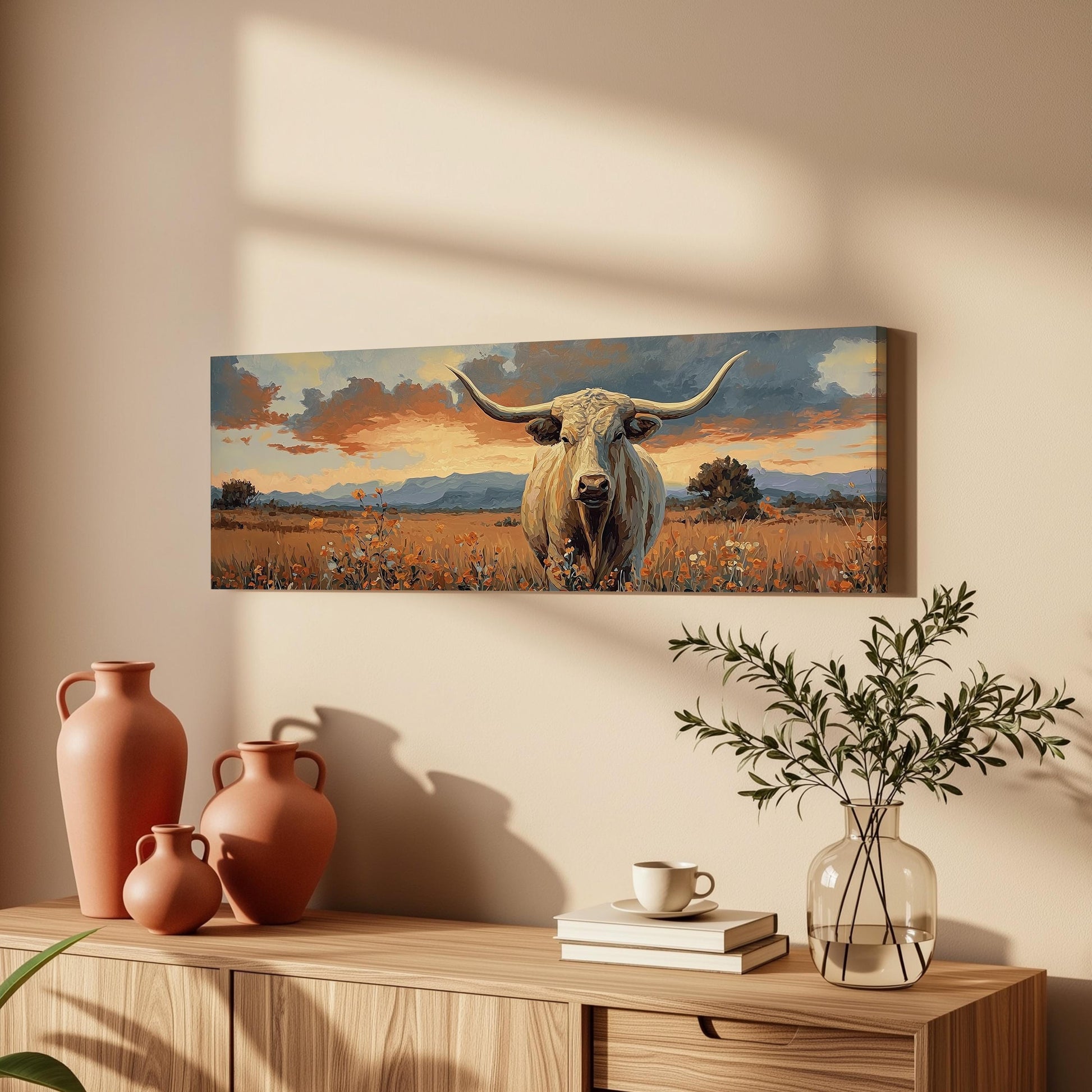 a large painting of a bull with horns, hanging on a wall above a wooden cabinet. The painting depicts a serene landscape with a field of flowers and a sunset sky. The cabinet has a vase, a coffee cup, and some books on it, creating a cozy and inviting atmosphere.