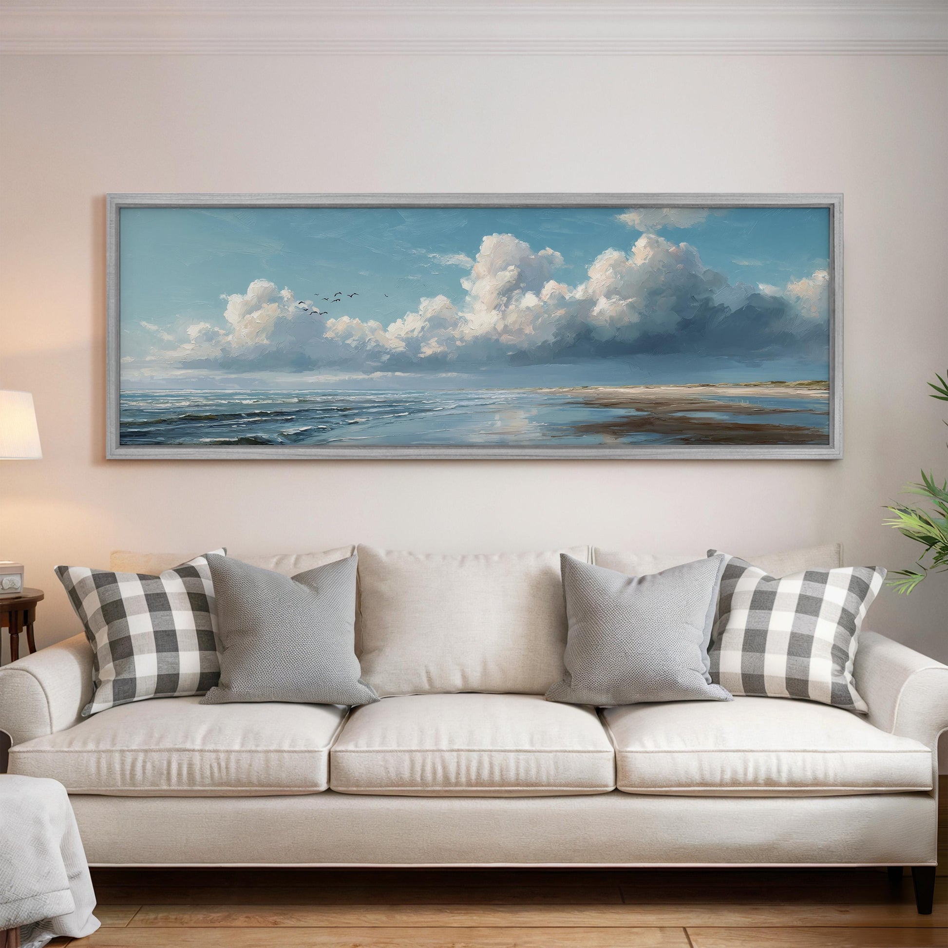 a large, framed painting of a serene landscape with a cloudy sky and ocean, hanging on a wall above a white sofa with gray and white patterned pillows.