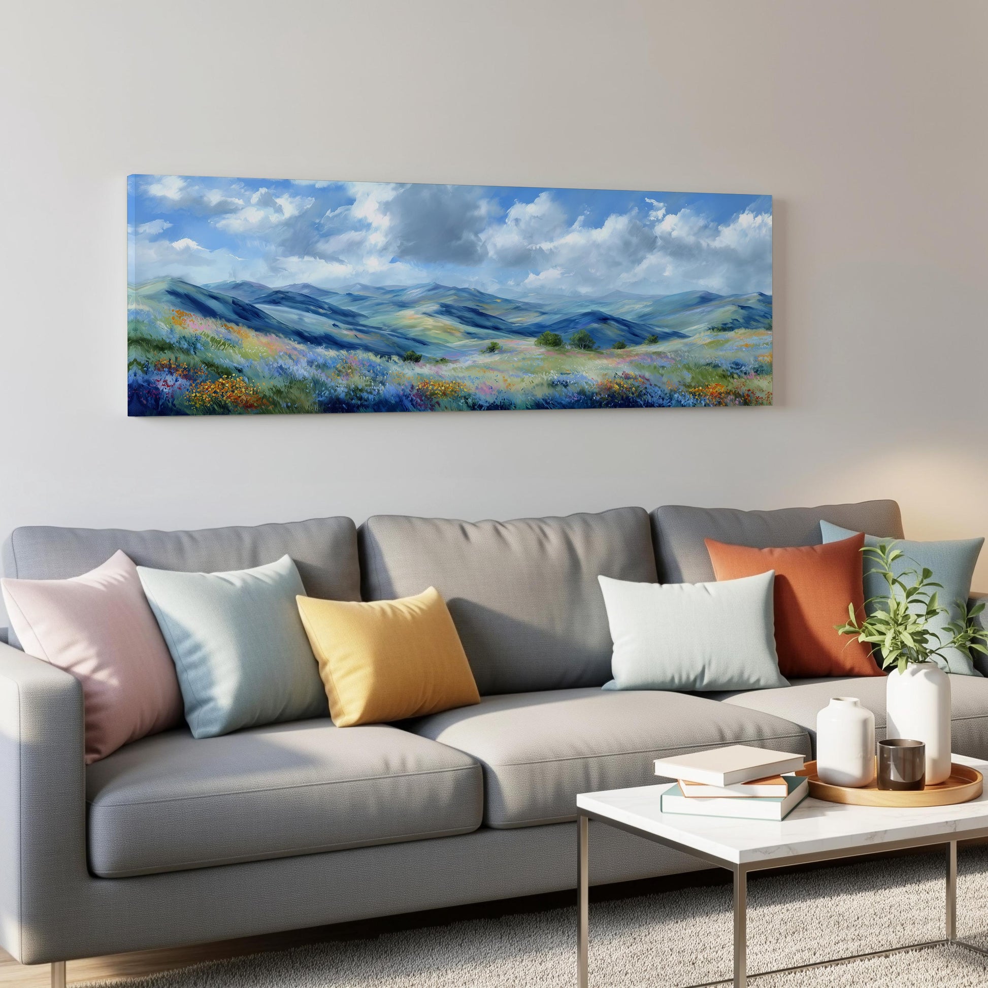 a large landscape painting hanging on a wall above a gray couch with colorful throw pillows. The painting depicts a serene countryside scene with rolling hills, a blue sky with clouds, and a field of flowers. The couch is positioned in front of the painting, creating a cozy and inviting atmosphere in the living room.