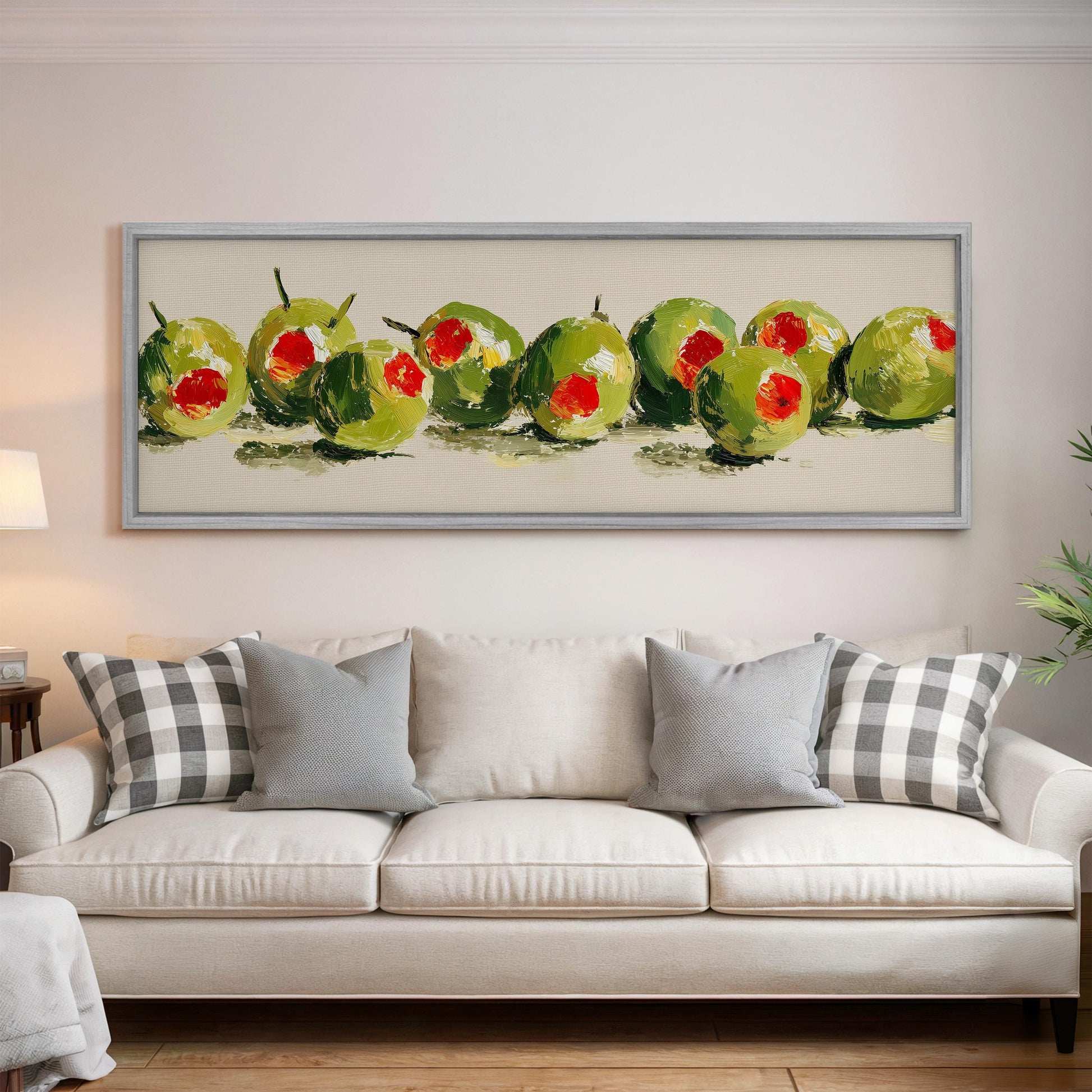 a large framed painting of green olives hanging on a wall above a white couch with gray and white plaid throw pillows.