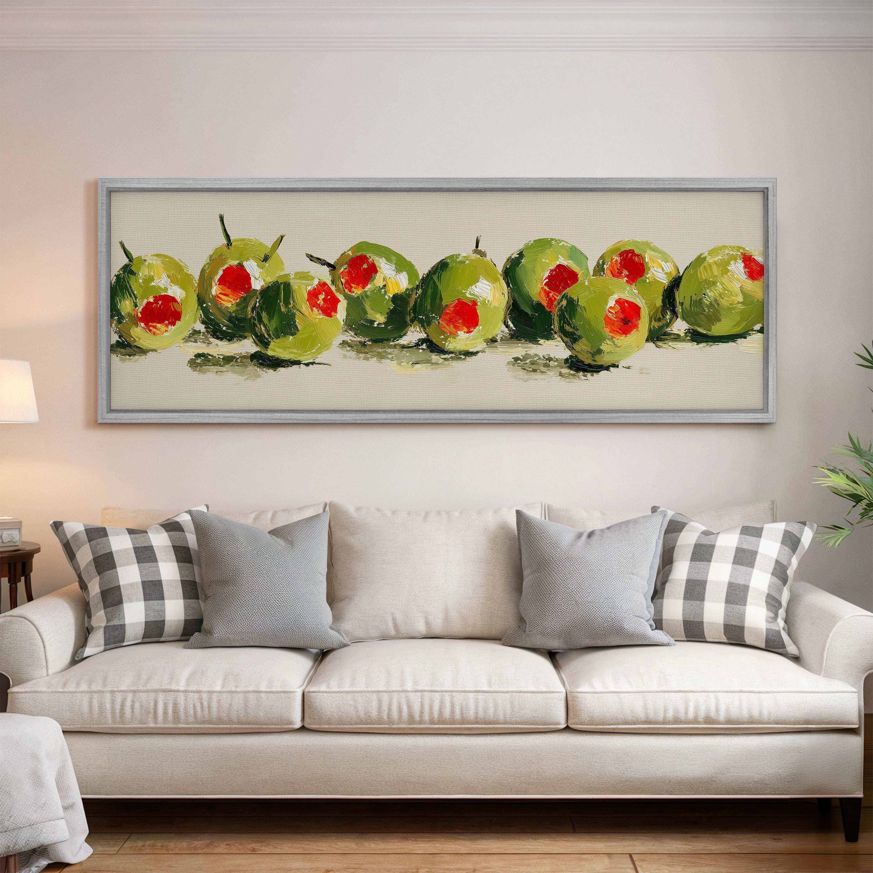 a large framed painting of green olives hanging on a wall above a white couch with gray and white plaid throw pillows.