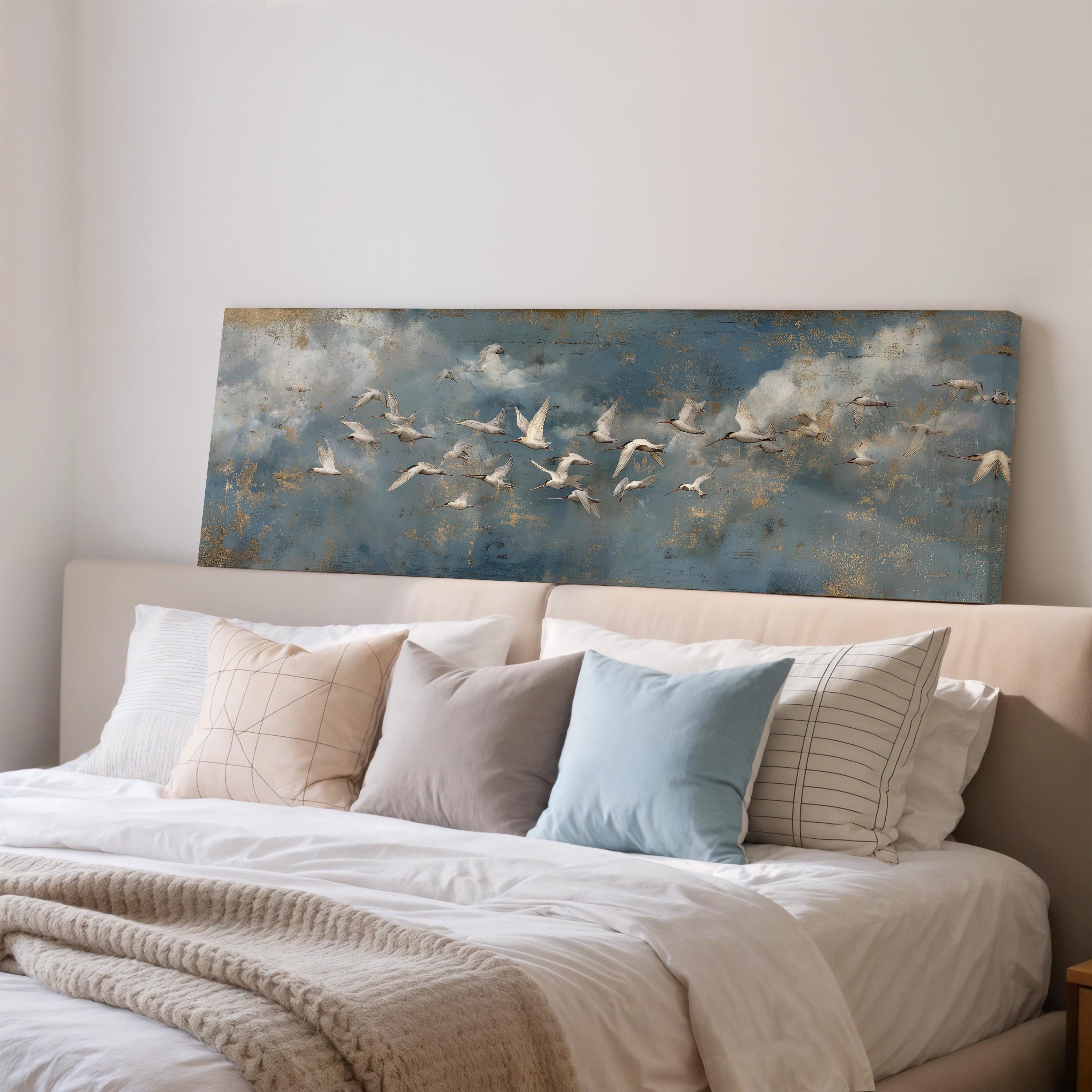 a bedroom with a large bed covered in white sheets and pillows. Above the bed, there is a painting of a flock of white birds flying across a blue sky with clouds.