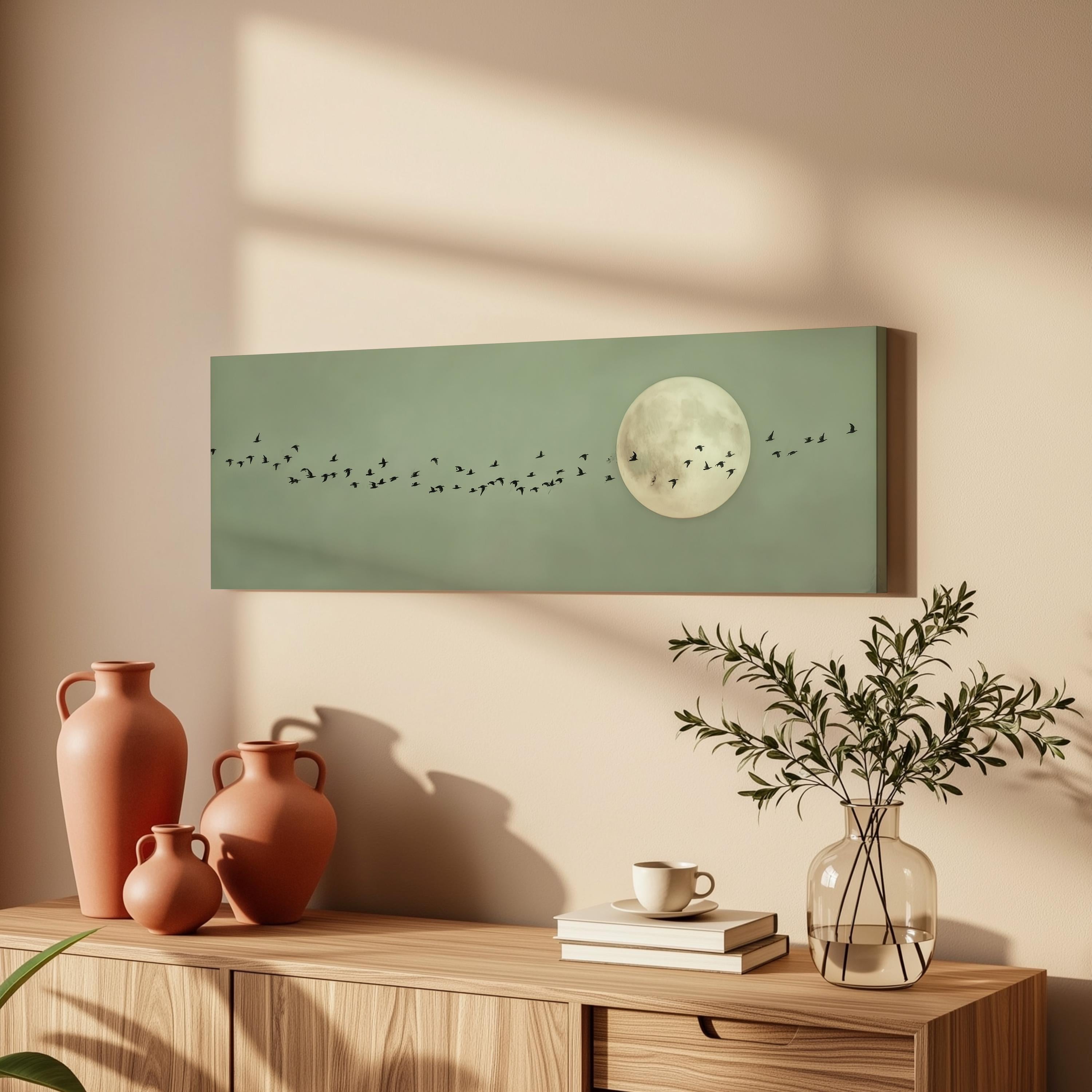 a wooden dresser with a vase of greenery, a coffee cup, and a stack of books. Above the dresser, there is a framed artwork depicting a flock of birds flying in front of a full moon.