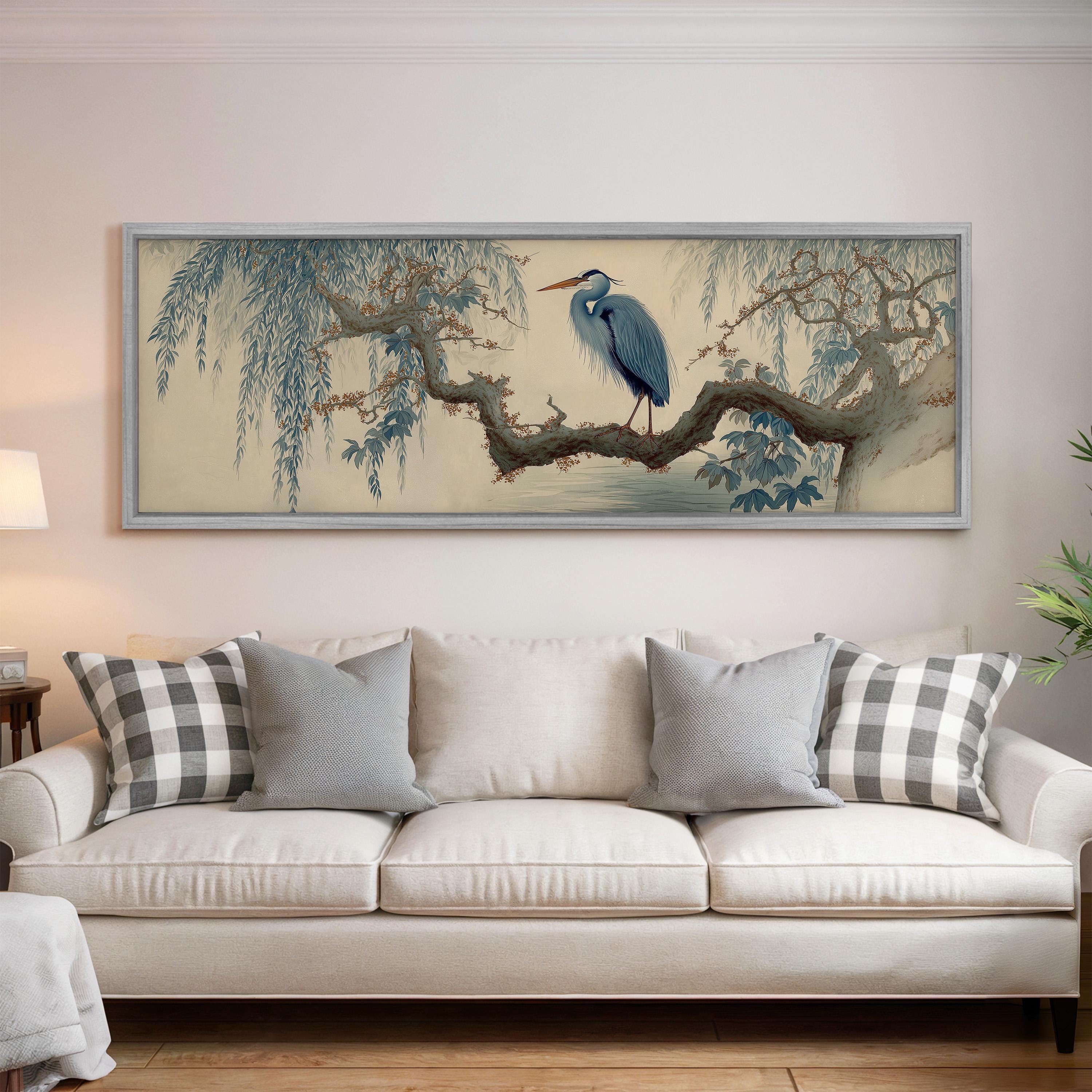 a large, framed painting of a blue heron perched on a branch, hanging on a wall above a white couch with gray and white plaid throw pillows.