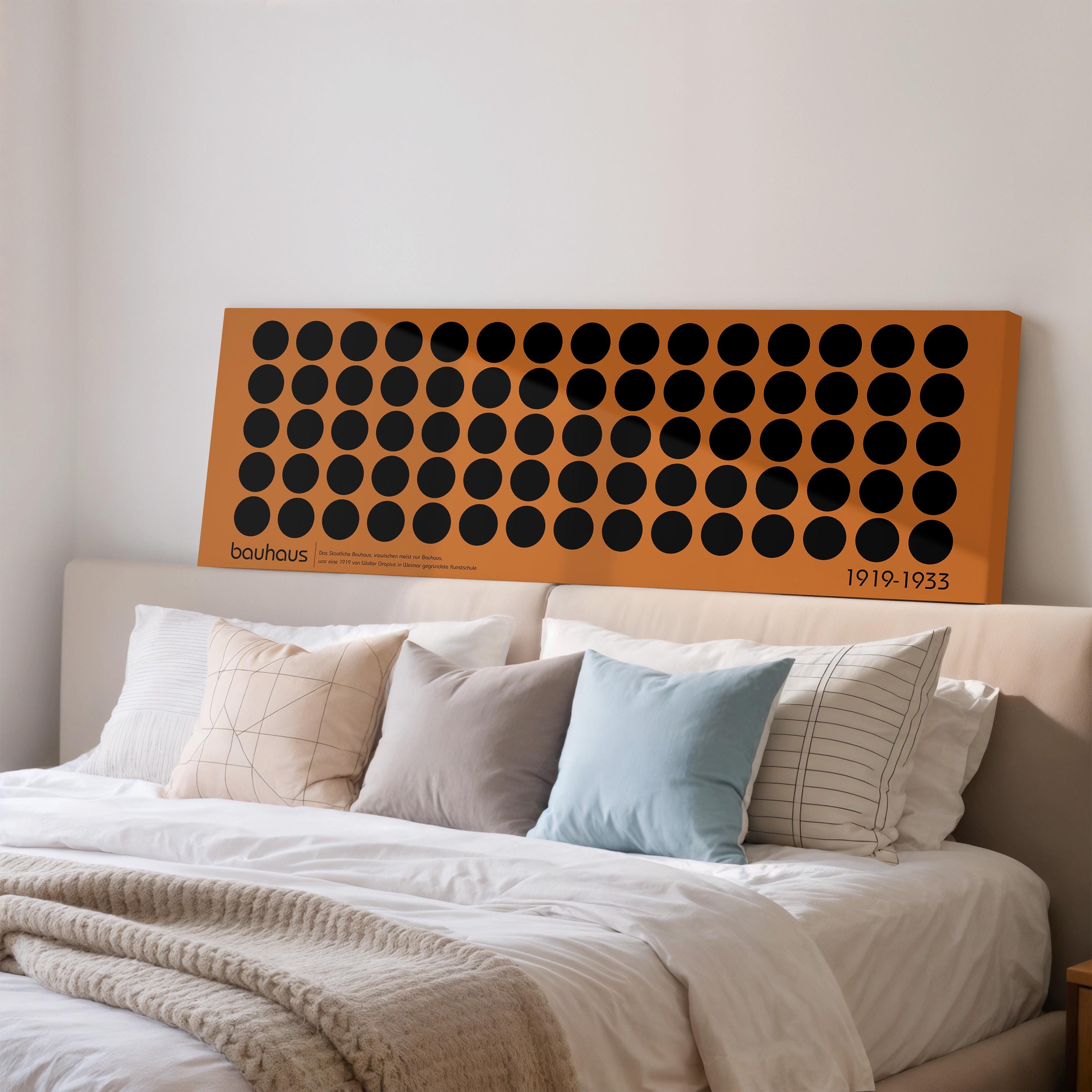 a large orange canvas with black circles on it, which is mounted on a white wall above a bed with white pillows and a gray blanket.
