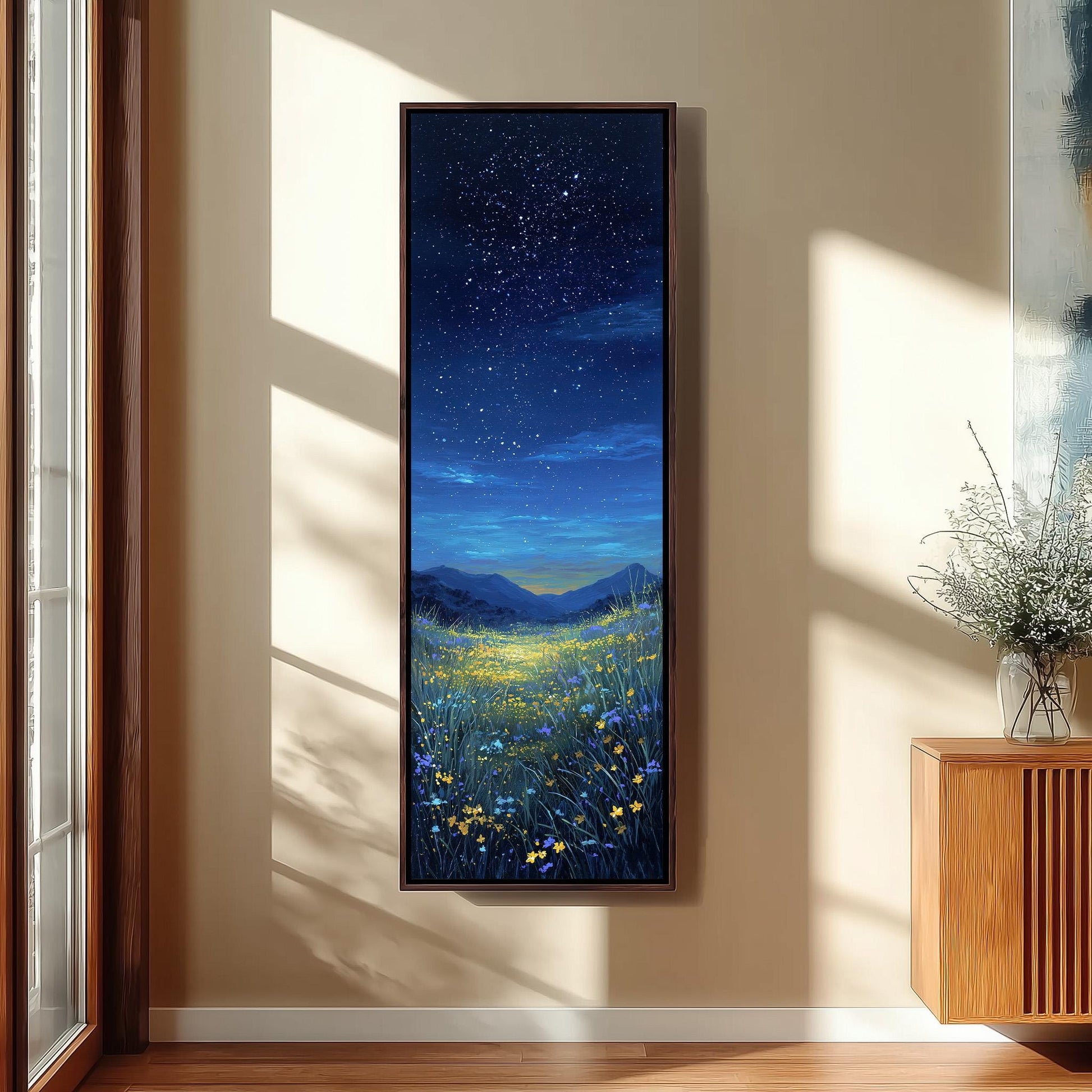 a large framed painting of a night sky with stars and a field of flowers, hanging on a wall in a room with a wooden cabinet and a vase of flowers.