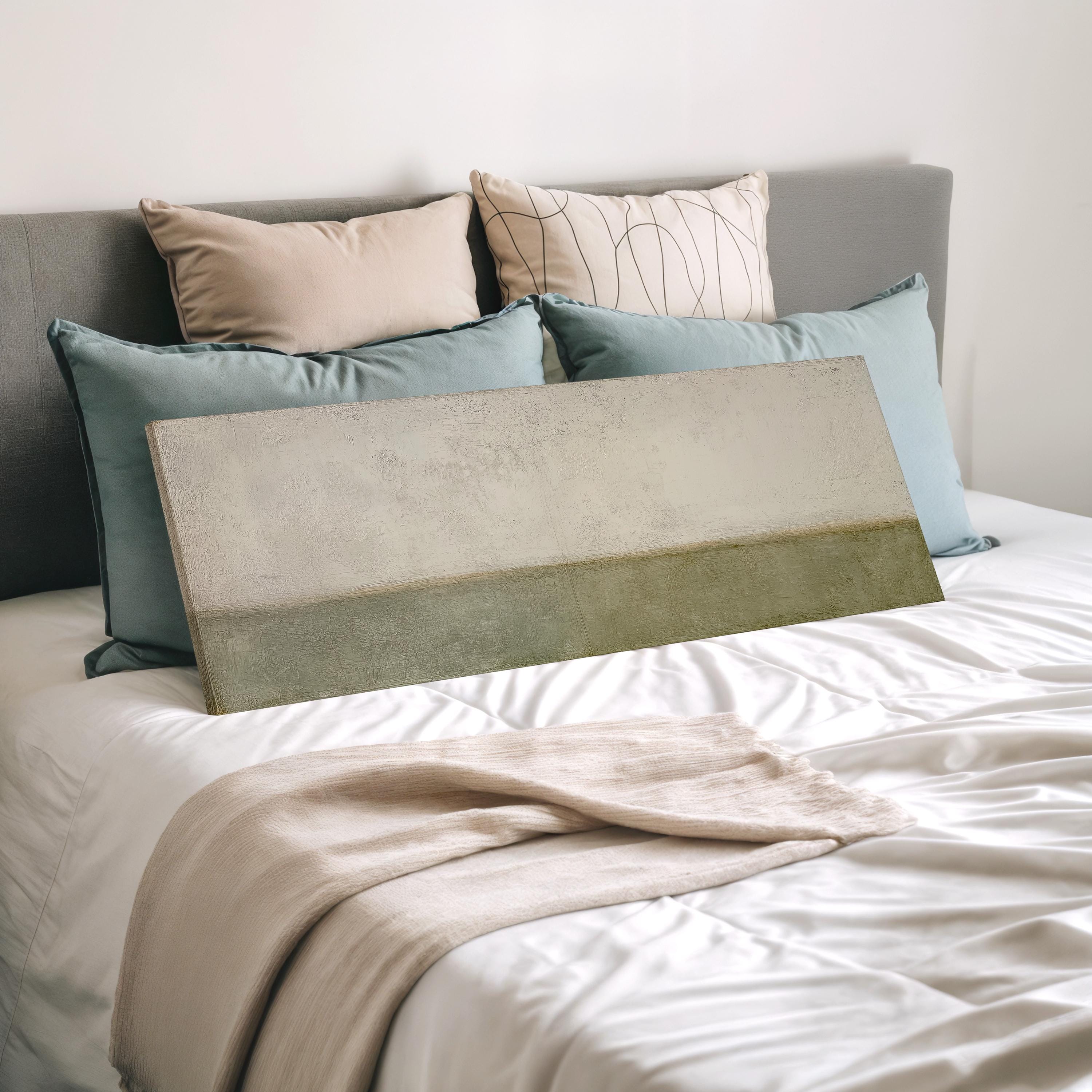 a neatly made bed with a white comforter and a gray headboard. The bed is adorned with a beige blanket and a gray throw pillow. There is a large rectangular painting or canvas resting on the bed, which appears to be a piece of art or a decorative item.