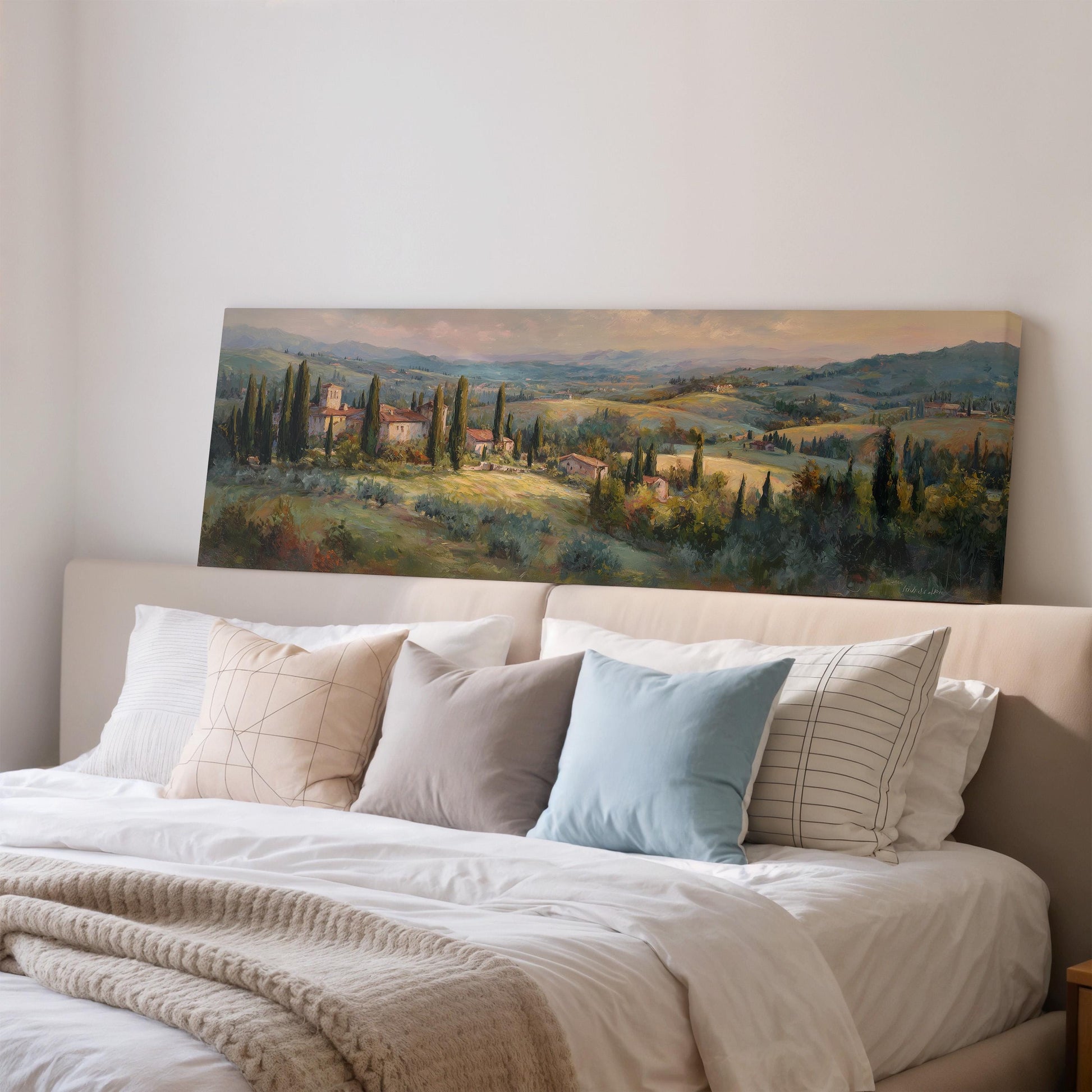a large painting of a countryside landscape hanging on a wall above a bed with white pillows and a gray blanket.