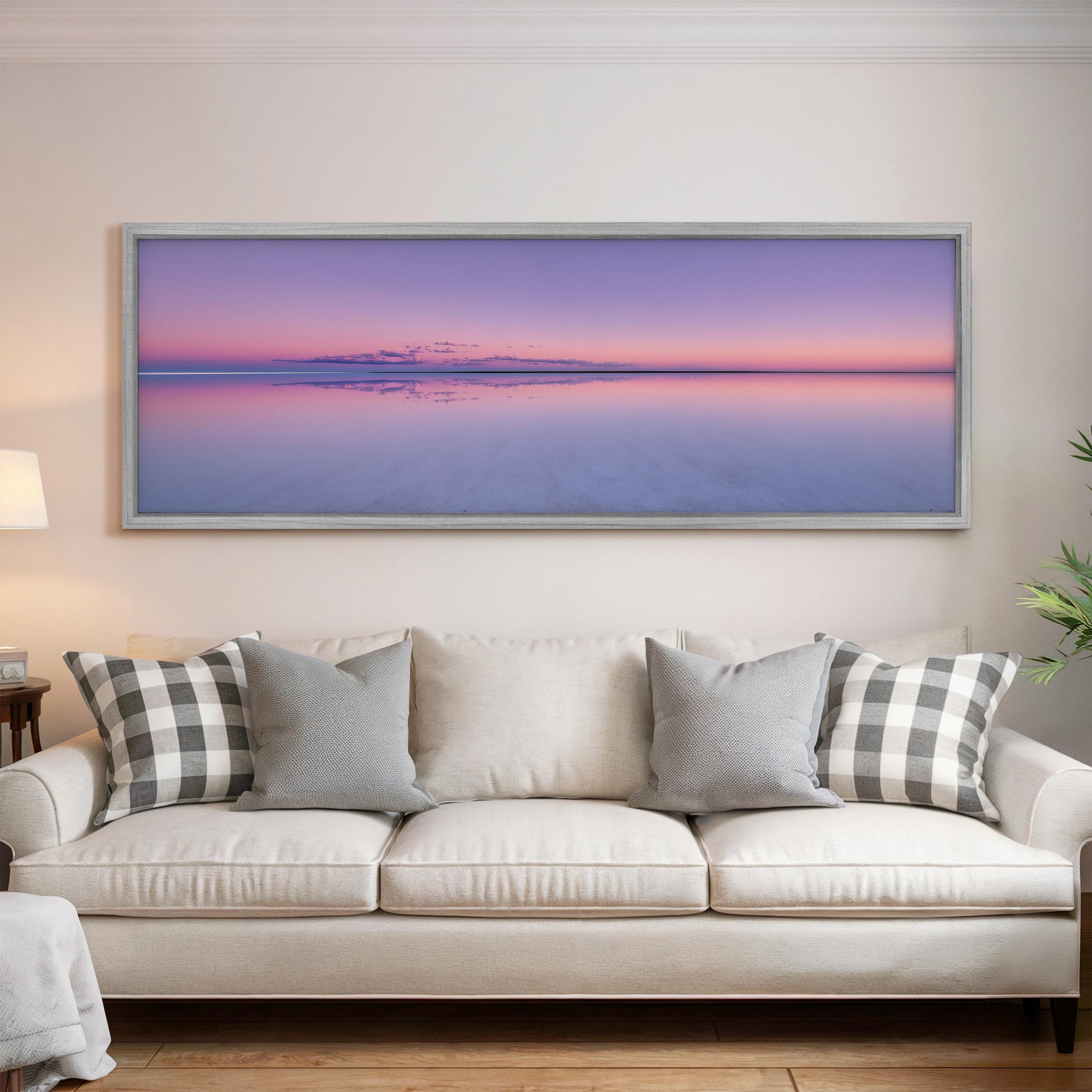 a large, framed picture of a serene landscape with a calm body of water reflecting the colors of the sky, including shades of pink and purple. The picture is hung on a wall above a white couch with gray and white patterned pillows.