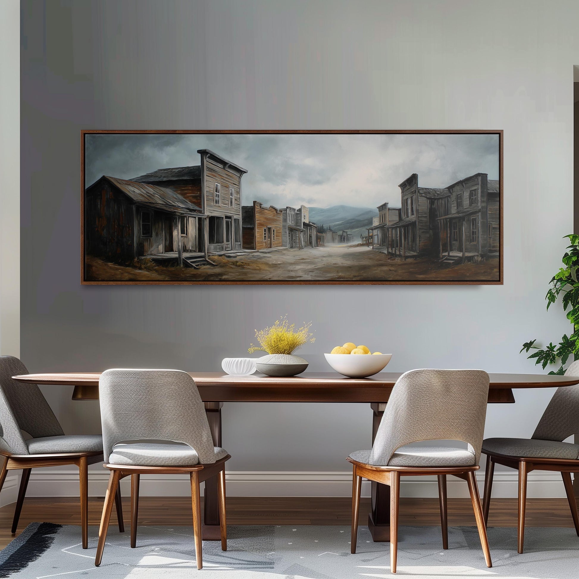 a dining room with a large painting of a town scene hanging on the wall above a wooden table with four chairs. The painting depicts a quaint, old-fashioned town with buildings, a road, and a cloudy sky. The dining table is set with a bowl of fruit and a vase of flowers, creating a cozy and inviting atmosphere.