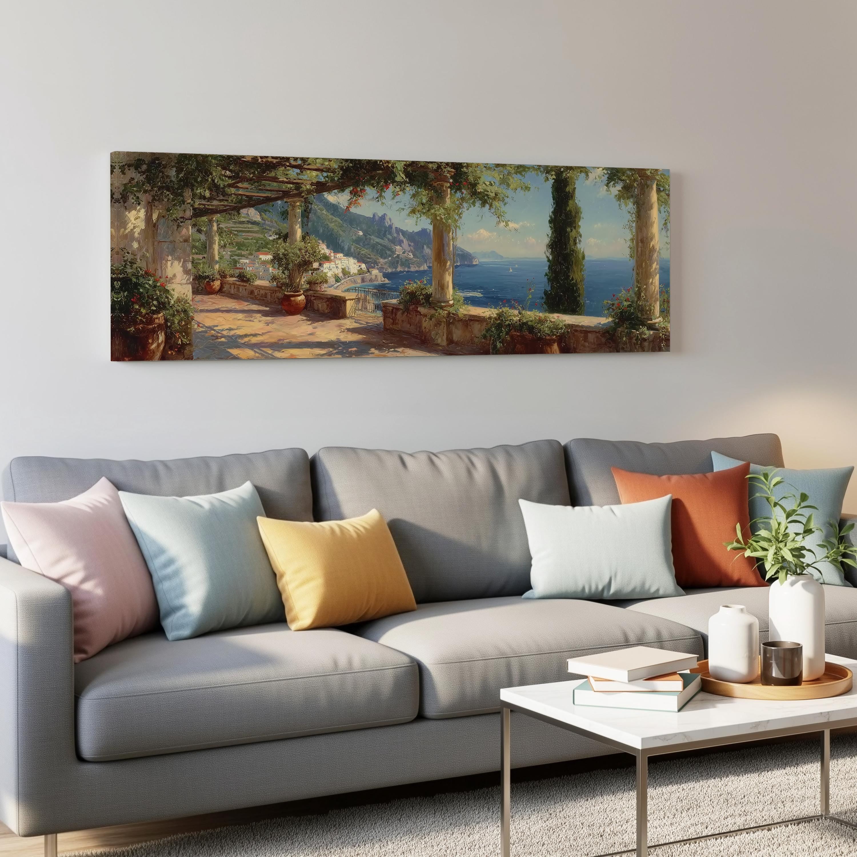 a living room with a gray sofa adorned with colorful throw pillows, a coffee table with books and a plant, and a large painting of a coastal landscape hanging on the wall above the sofa.