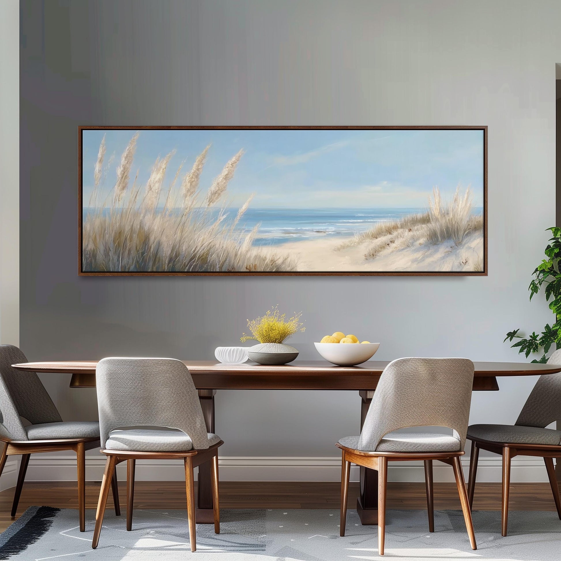 a dining room with a large painting of a beach scene hanging on the wall above a wooden dining table with four chairs. The painting depicts a sandy beach with tall grasses and a blue sky with clouds. On the table, there is a bowl of fruit and a vase of flowers, adding a touch of color and life to the room.