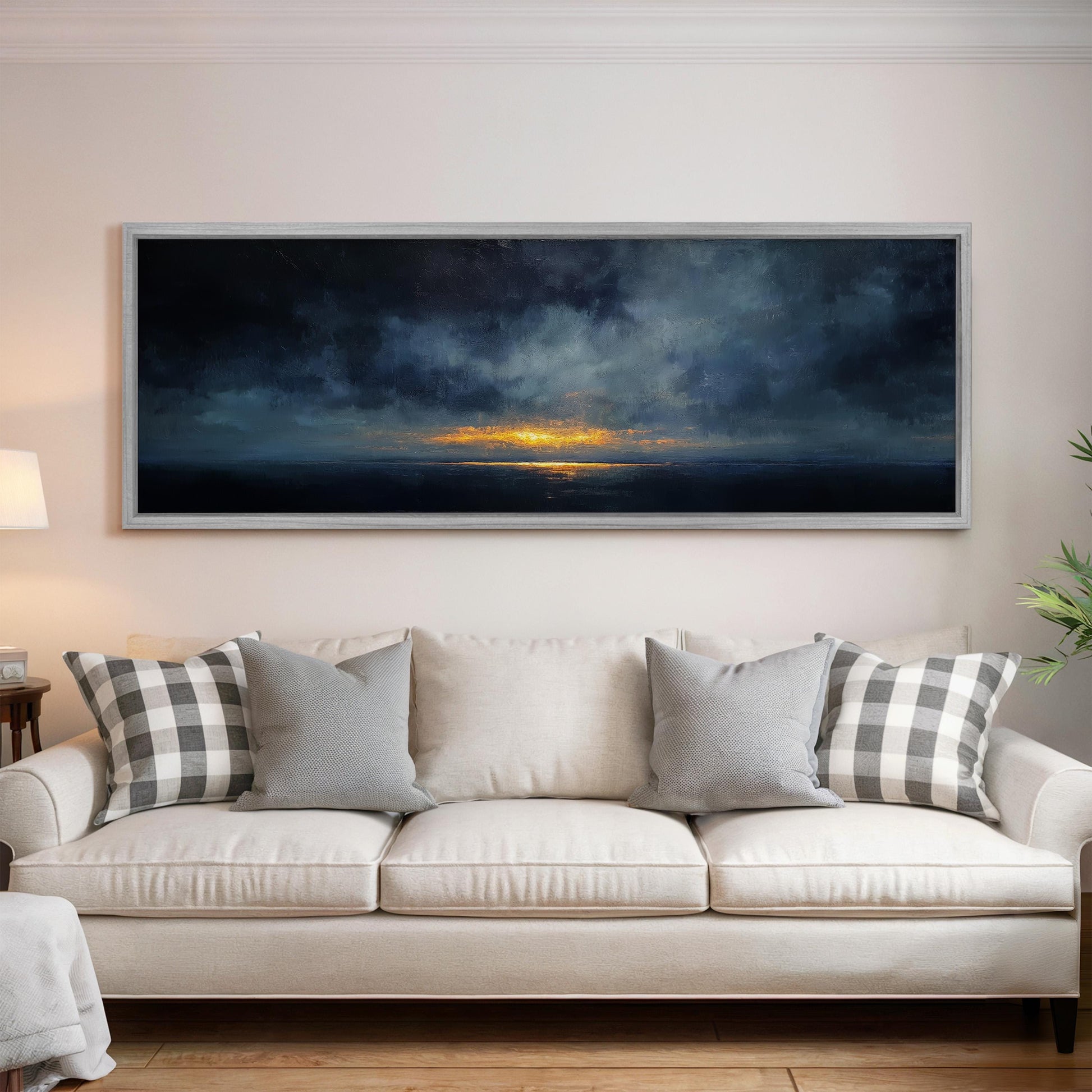 a large, dark-toned painting of a sunset over the ocean, hanging on a wall above a white sofa with gray and white patterned throw pillows.