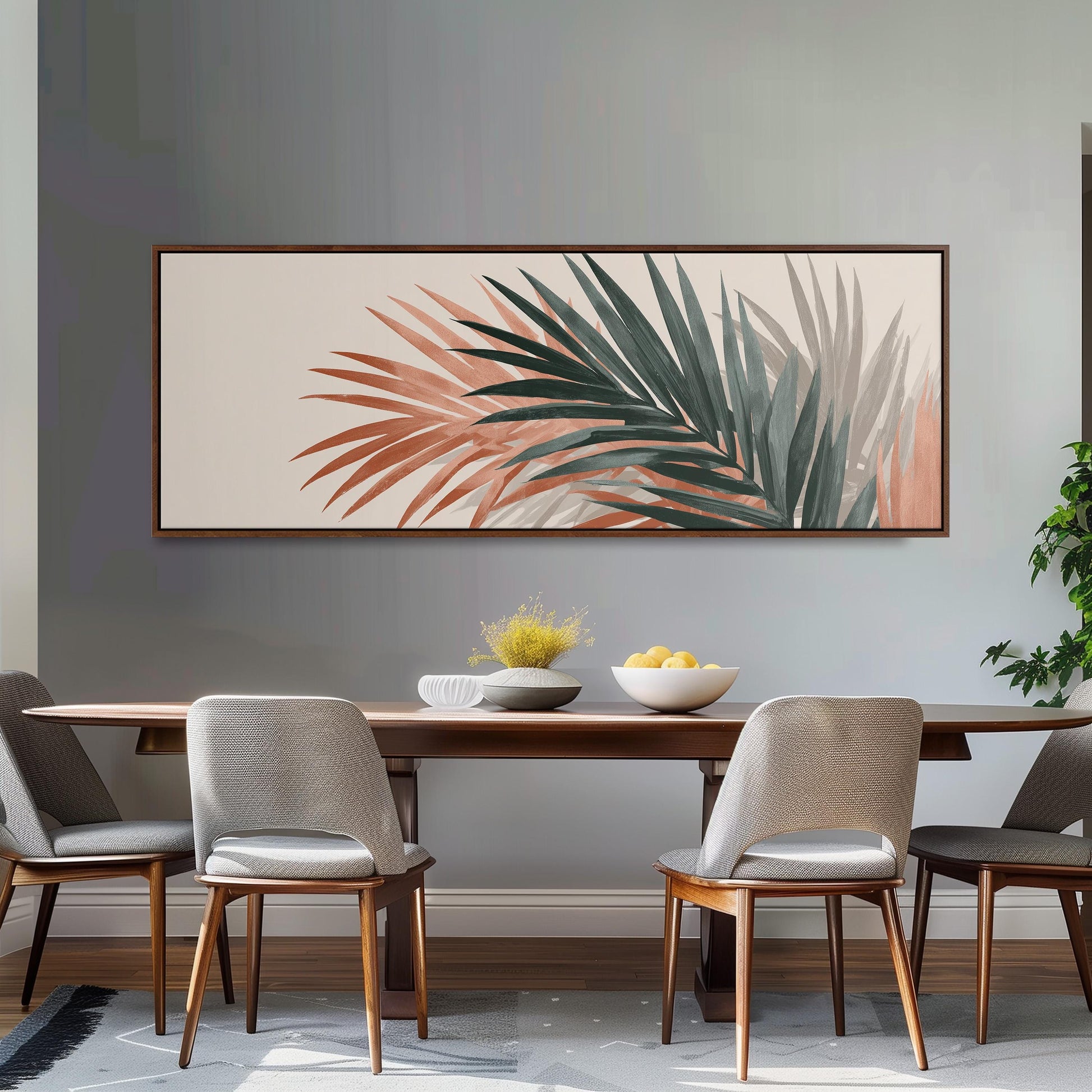 a dining room with a wooden table and four chairs. Above the table, there is a large framed artwork depicting palm leaves in various shades of green and orange. The artwork is hung on a gray wall above the table. The table is set with a bowl of fruit and a vase of flowers, creating a cozy and inviting atmosphere.