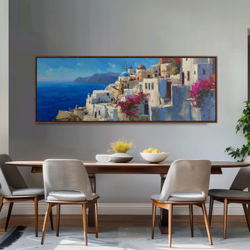 a large painting of a coastal town with white buildings and pink flowers hanging above a wooden dining table with four chairs.