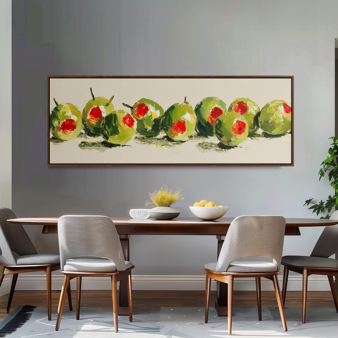 a dining room with a wooden table and four chairs. Above the table, there is a large painting of green olives with red centers. The painting is framed and hangs on the wall. The table is set with a bowl of fruit and a vase of flowers, creating a welcoming and inviting atmosphere.