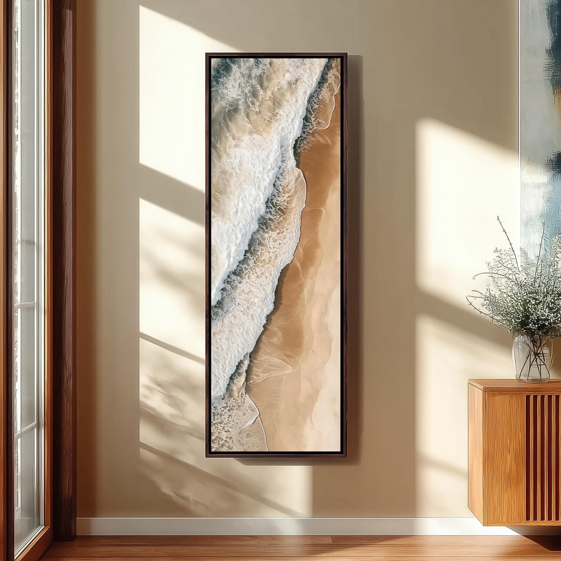 a large framed picture of a beach scene hanging on a wall, with a wooden cabinet and a vase of flowers to the right of the frame.