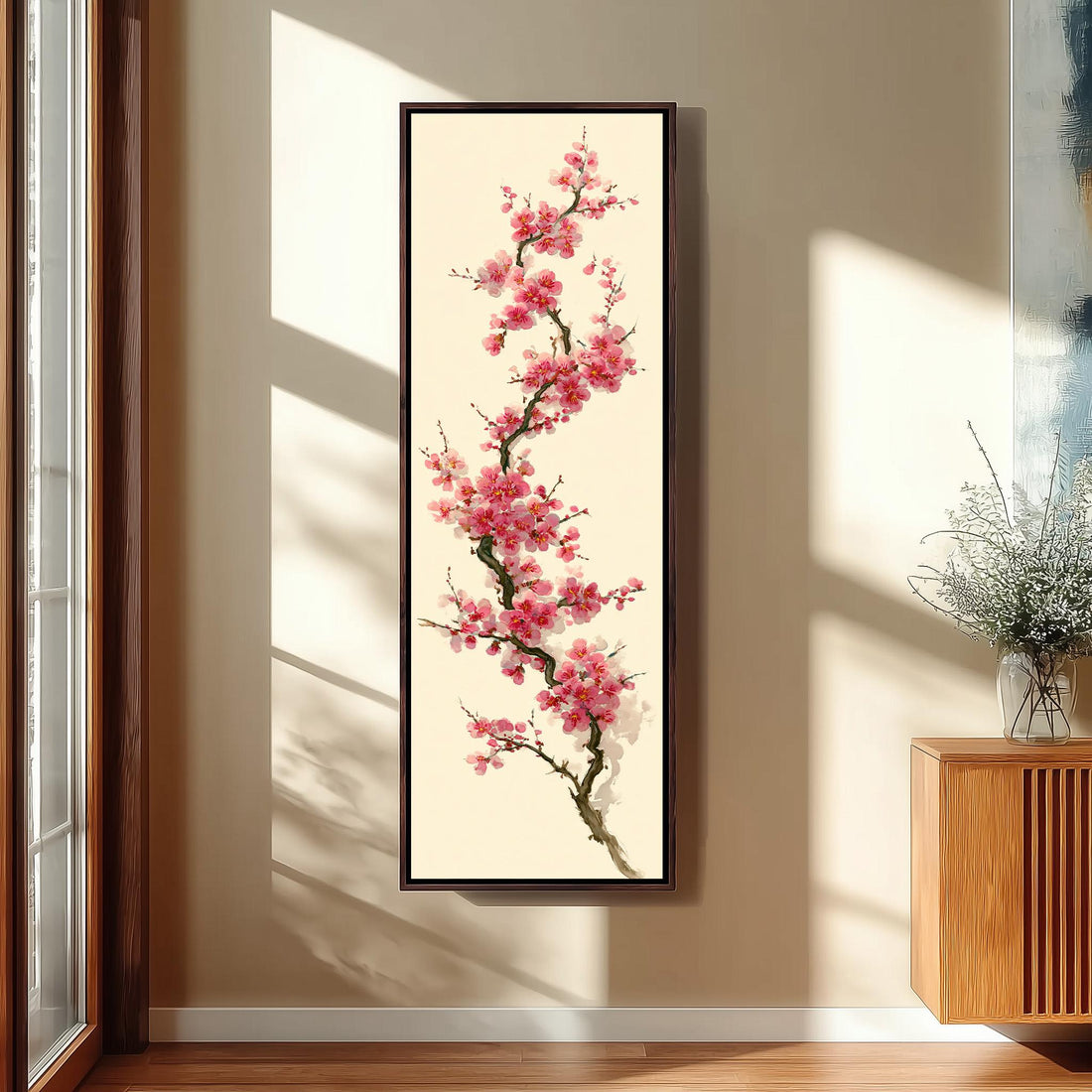 a large framed painting of a pink flower on a wall, with a wooden cabinet and a vase of flowers to the right of the painting.