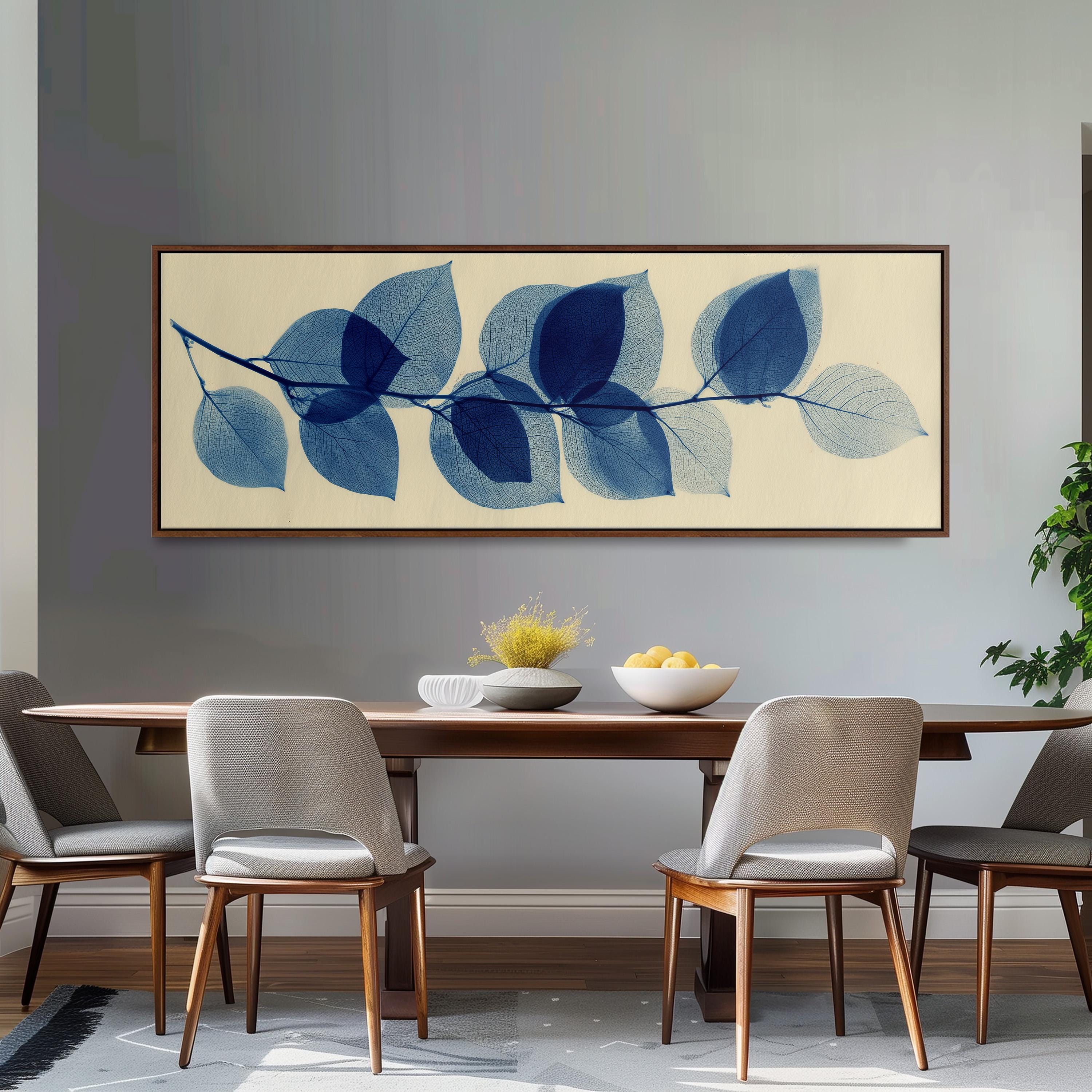a dining room with a wooden table and four chairs. Above the table, there is a large framed artwork depicting blue leaves on a branch. The table is set with a bowl of fruit and a vase of flowers.