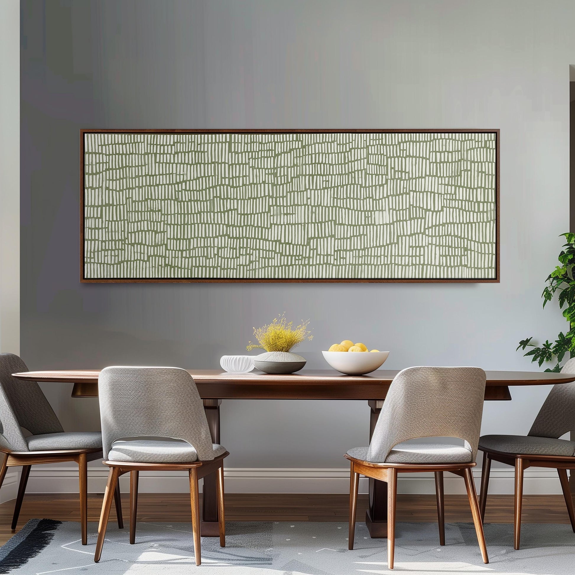 a modern dining room with a wooden table and four gray chairs. Above the table, there is a large abstract painting with a geometric pattern in shades of green and white. On the table, there is a bowl of fruit and a vase of flowers, adding a touch of color and life to the space.