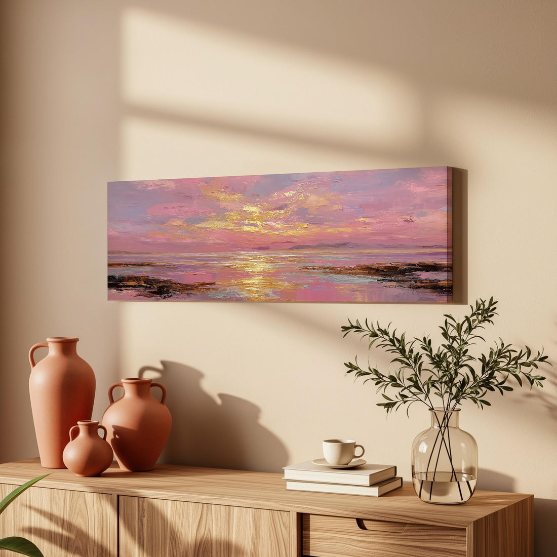 a wooden cabinet with a painting of a sunset landscape hanging on the wall above it. The painting is vibrant and colorful, depicting a serene sunset over a body of water. The cabinet also has three orange vases and a glass vase with greenery, as well as a stack of books and a cup on the surface.