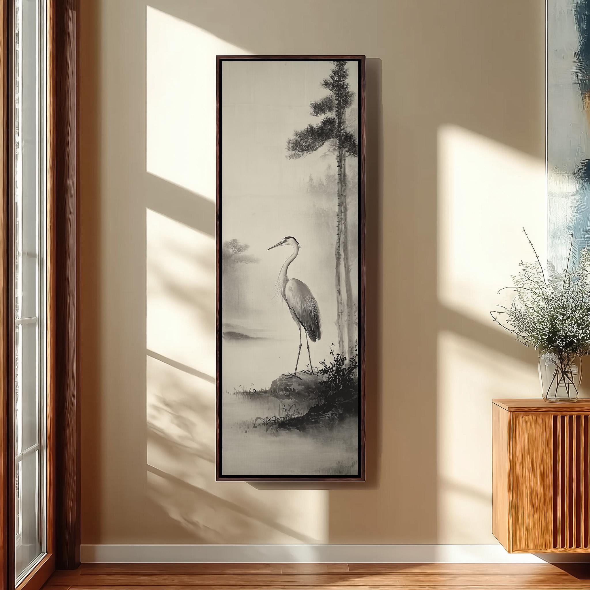 a large black and white painting of a crane standing on a rock, hanging on a wall in a room with a wooden cabinet and a vase of flowers.