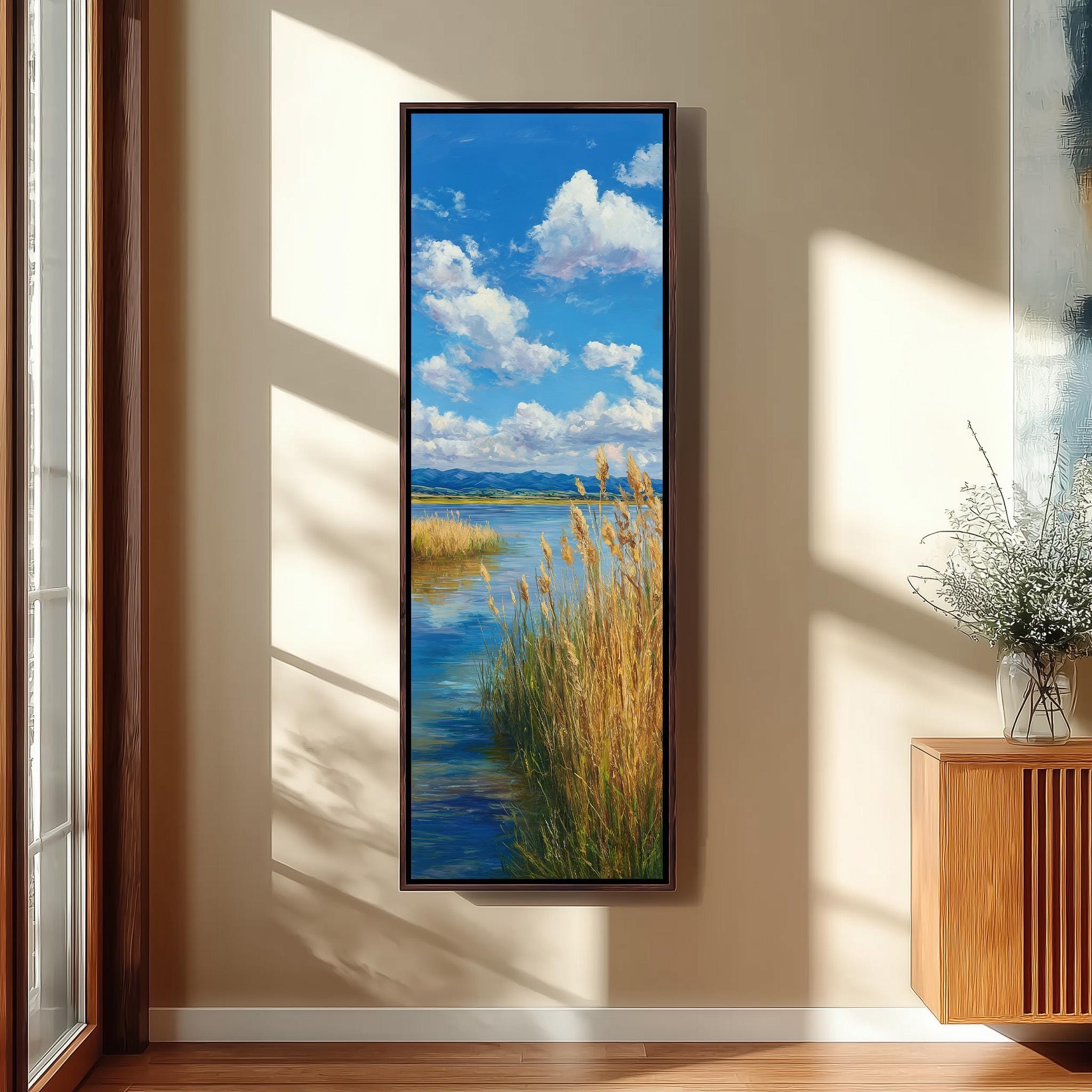 a large framed painting of a serene landscape with a river, grassy banks, and a blue sky with fluffy white clouds. The painting is hanging on a wall in a room with a wooden floor and a window.