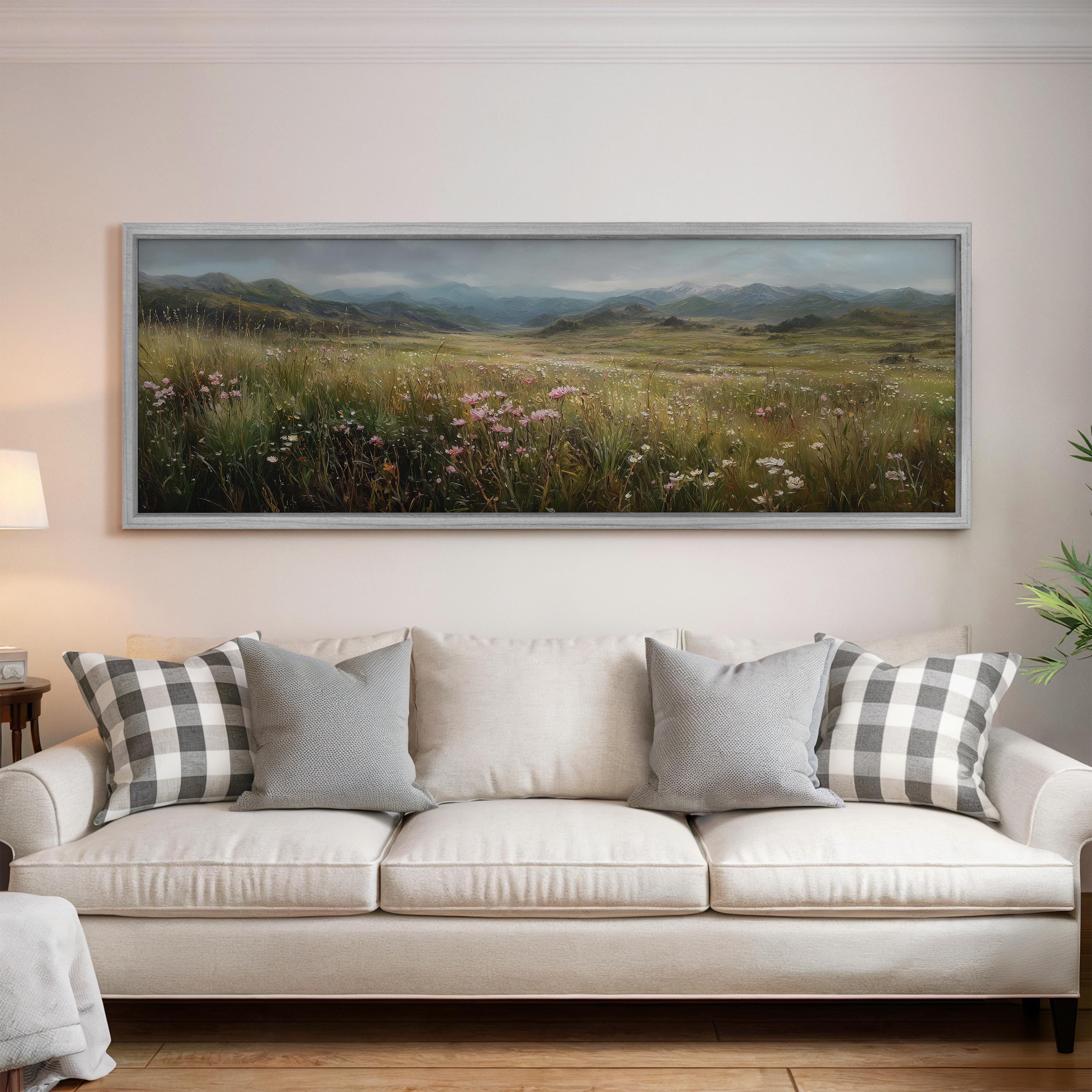a large, framed painting of a grassy field with wildflowers hanging on a wall above a white sofa with gray and white plaid throw pillows.