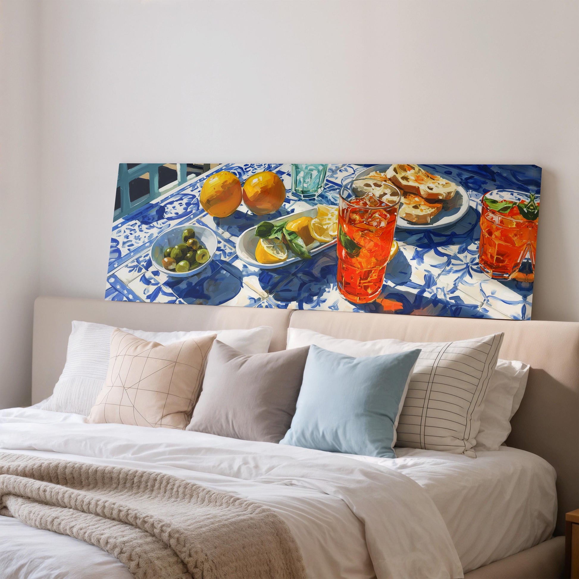 a large painting of a table with various food items, including oranges, a bowl of olives, and a glass of orange juice, displayed on a wall above a bed with white pillows and a beige blanket.