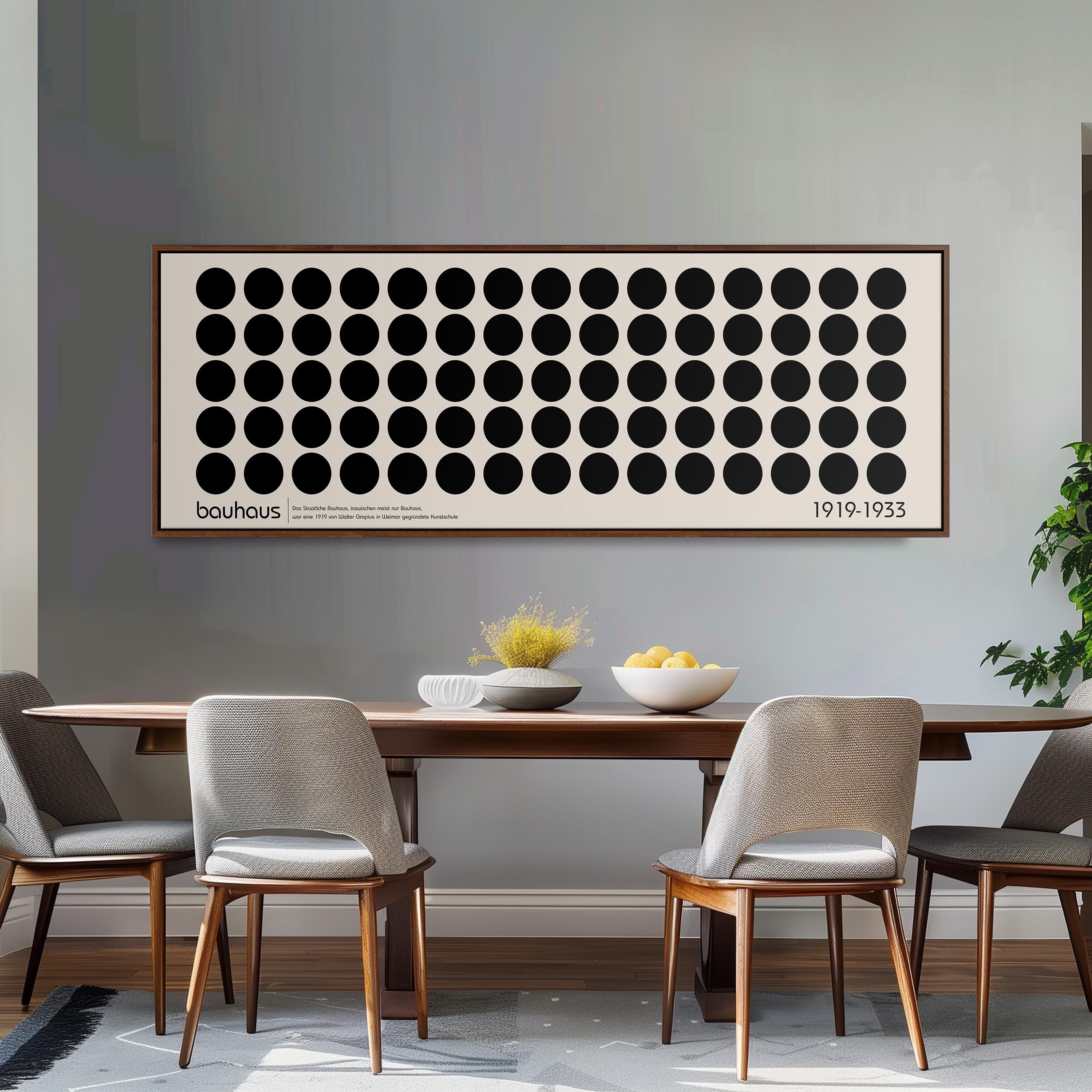 a dining room with a wooden table and four chairs. Above the table, there is a large framed artwork with a repeating pattern of circles. The artwork is framed in a white frame and is hung on a gray wall. The dining table is set with a bowl of fruit and a vase of flowers.