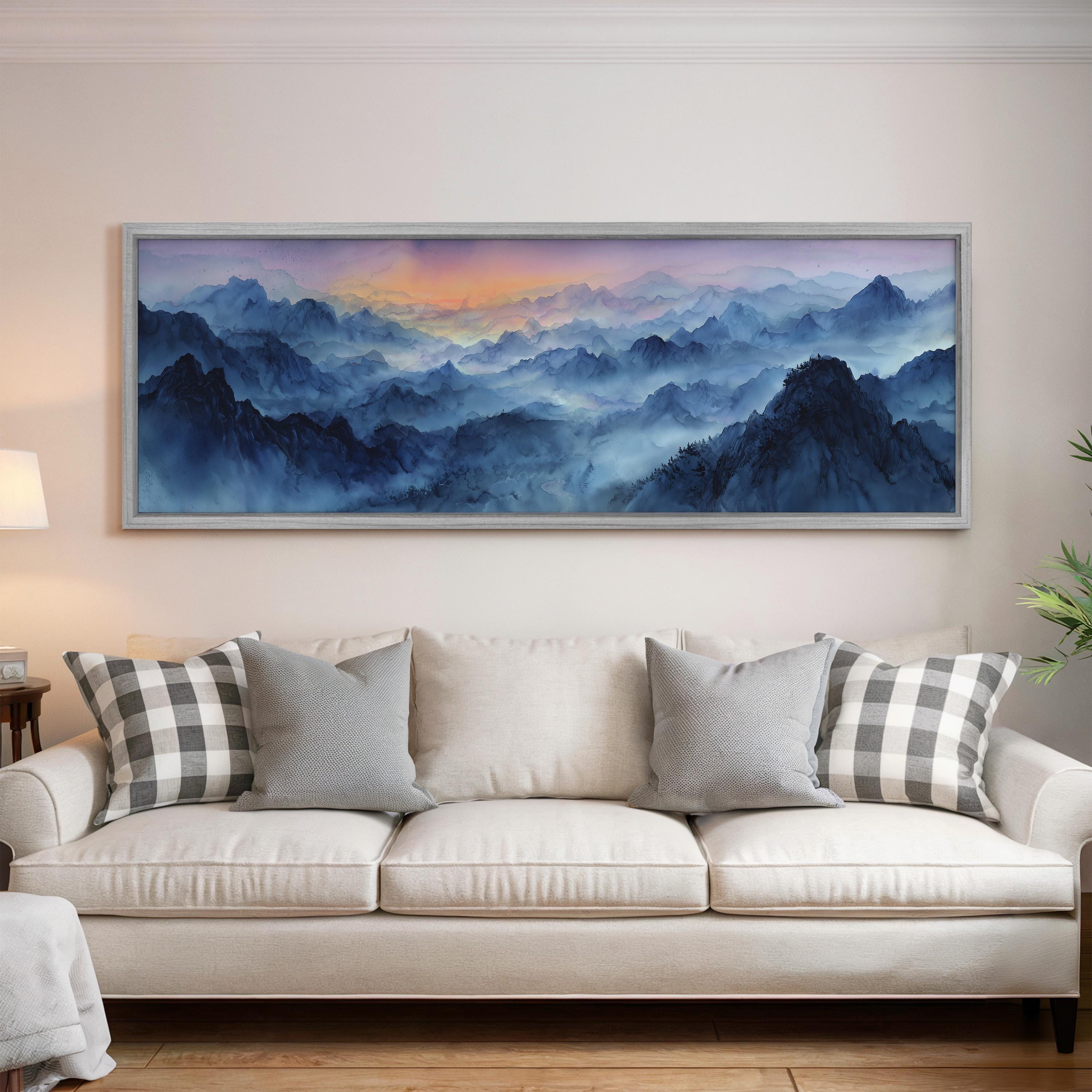 a large, framed painting of a mountainous landscape with a blue sky and orange and purple hues, hanging on a wall above a white couch with gray and white patterned pillows.