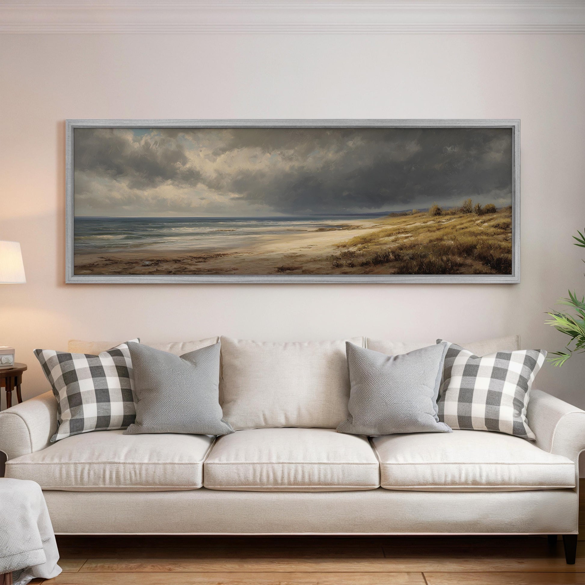 a large, framed painting of a beach scene hanging on a wall above a white sofa with gray and white patterned throw pillows.