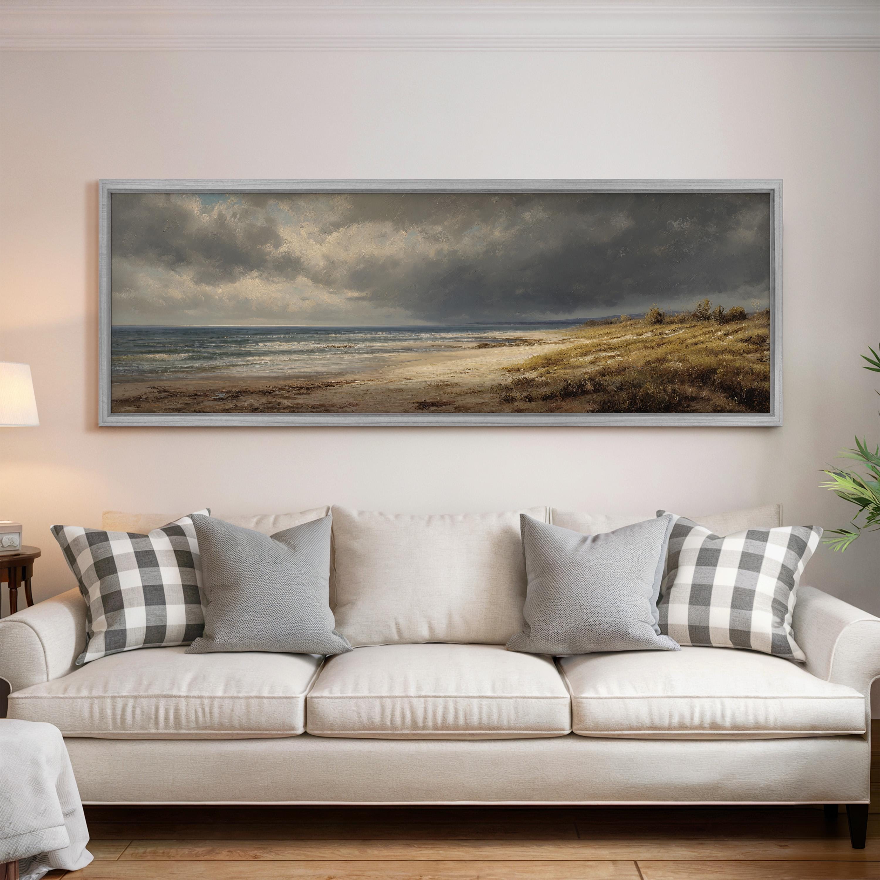 a large, framed painting of a beach scene hanging on a wall above a white sofa with gray and white patterned throw pillows.