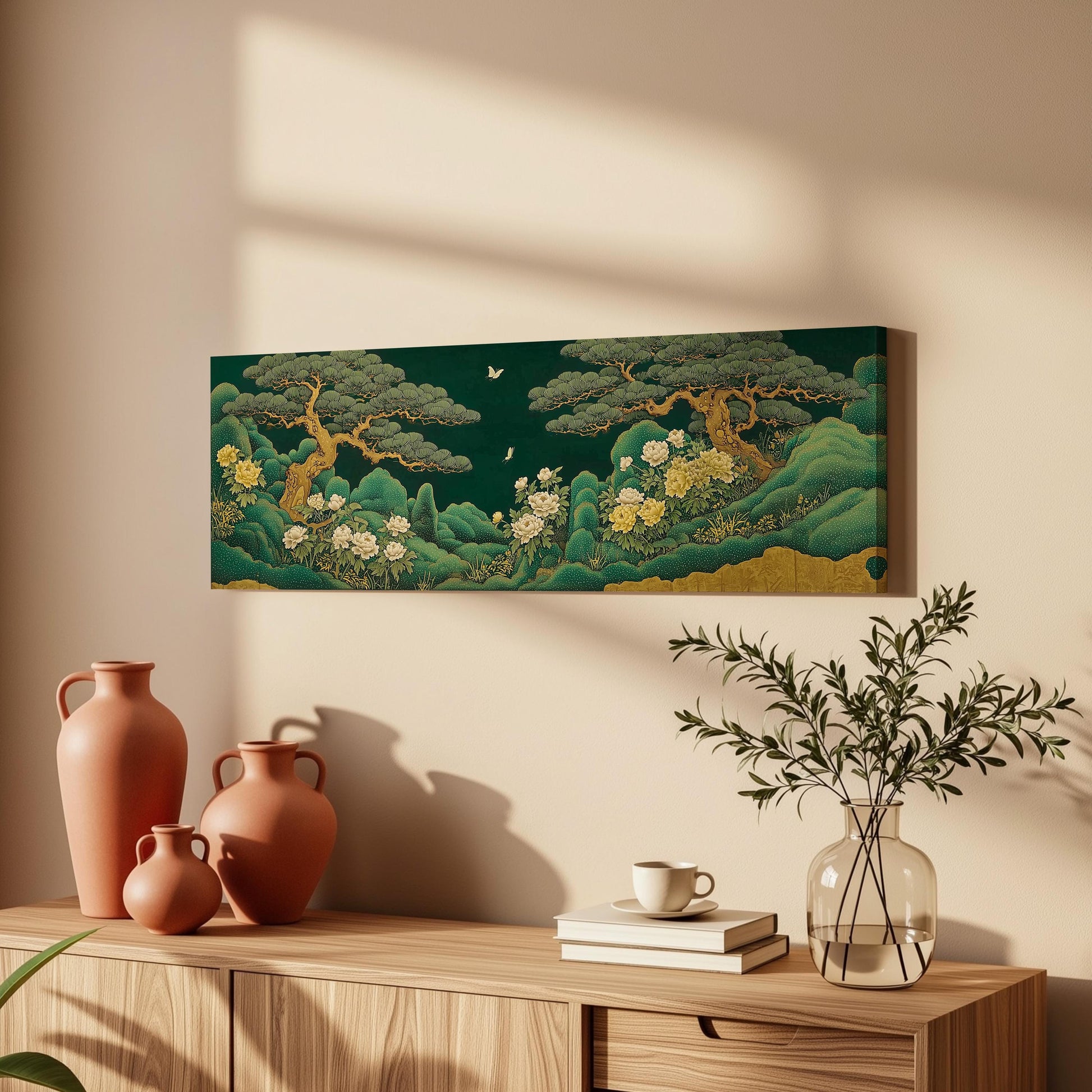 a wooden cabinet with a painting of a lush green landscape hanging on the wall above it. The cabinet also has three orange vases and a glass vase with greenery, as well as a stack of books and a cup on the surface.