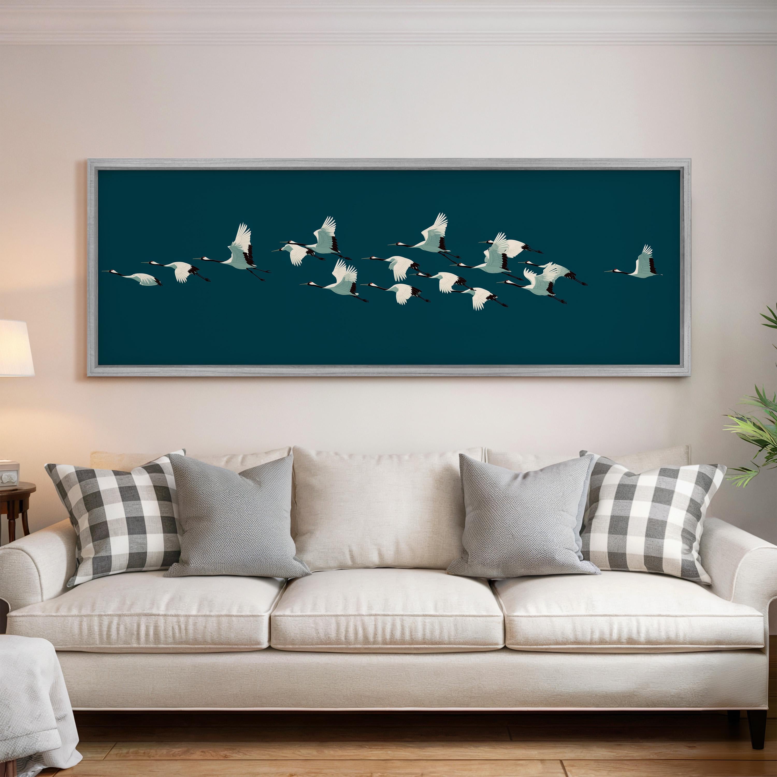 a large framed artwork of a flock of white birds flying across a dark blue background, hanging above a white couch with gray and white patterned pillows.