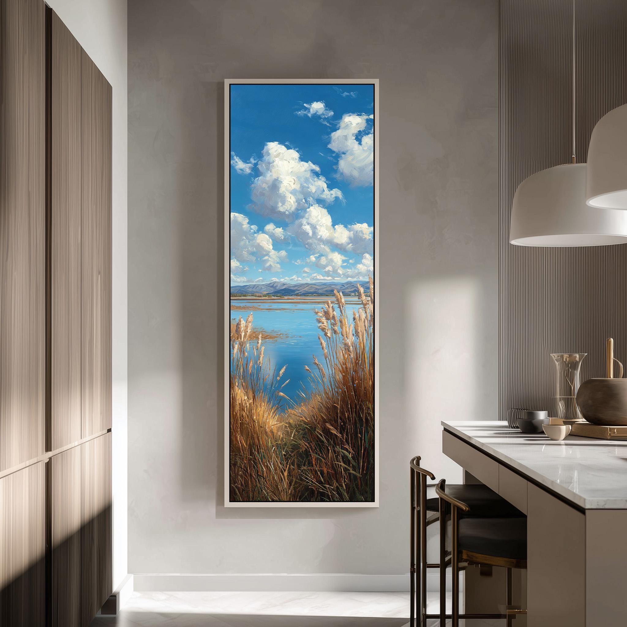 a large framed painting of a serene landscape with a lake, mountains, and a blue sky with fluffy white clouds. The painting is hung on a wall in a modern kitchen with a countertop and chairs.