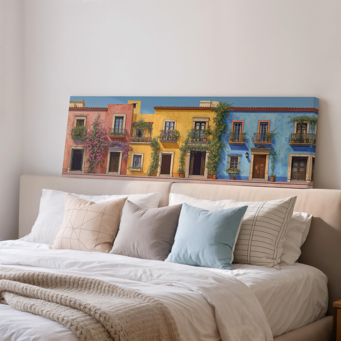 a wooden dresser with a vase of greenery, a stack of books, and a ceramic vase with flowers. Above the dresser, there is a painting of colorful buildings with balconies and a blue wall.