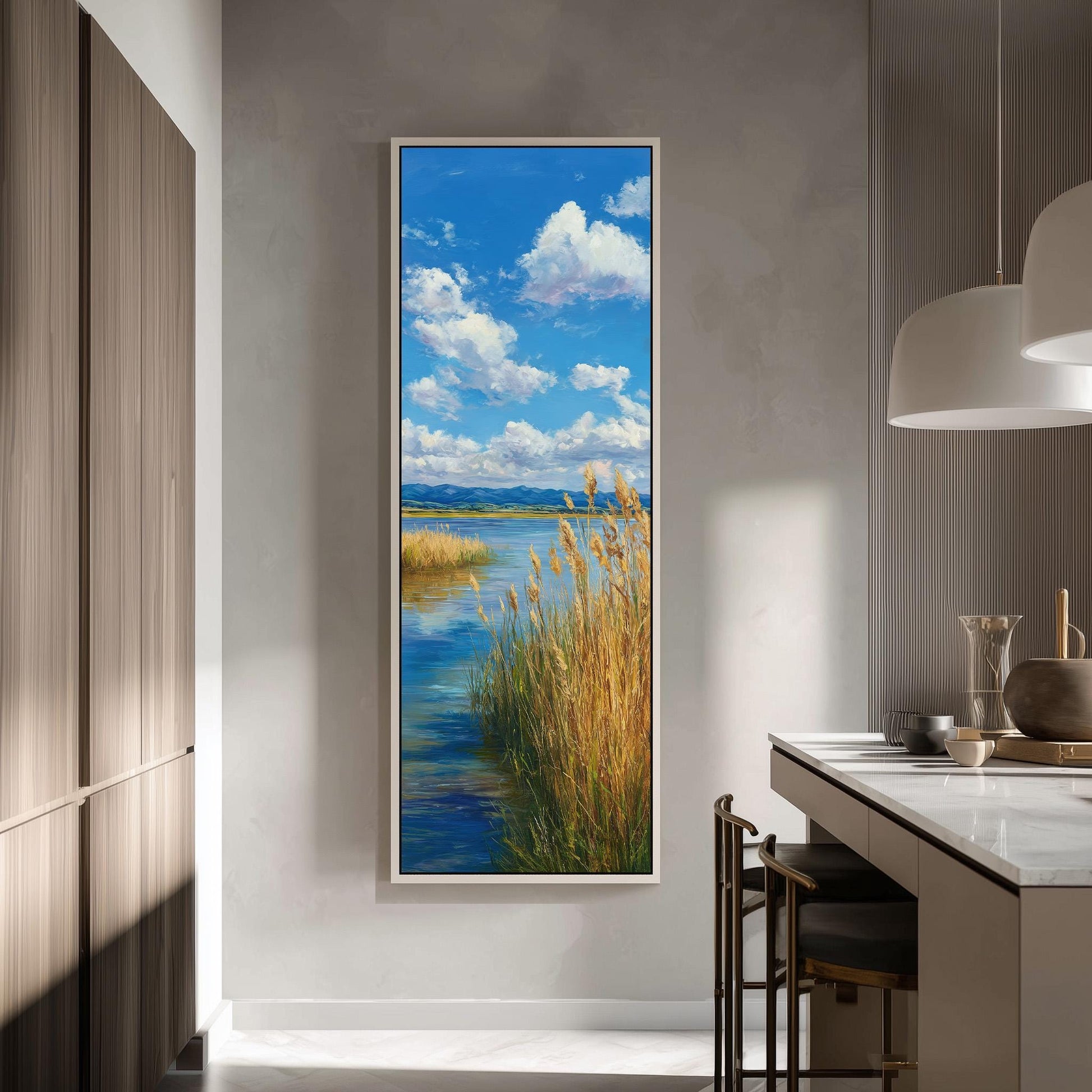 a large framed painting of a serene landscape with a river, grassy banks, and a blue sky with fluffy white clouds. The painting is hung on a wall in a modern kitchen with a countertop and chairs.