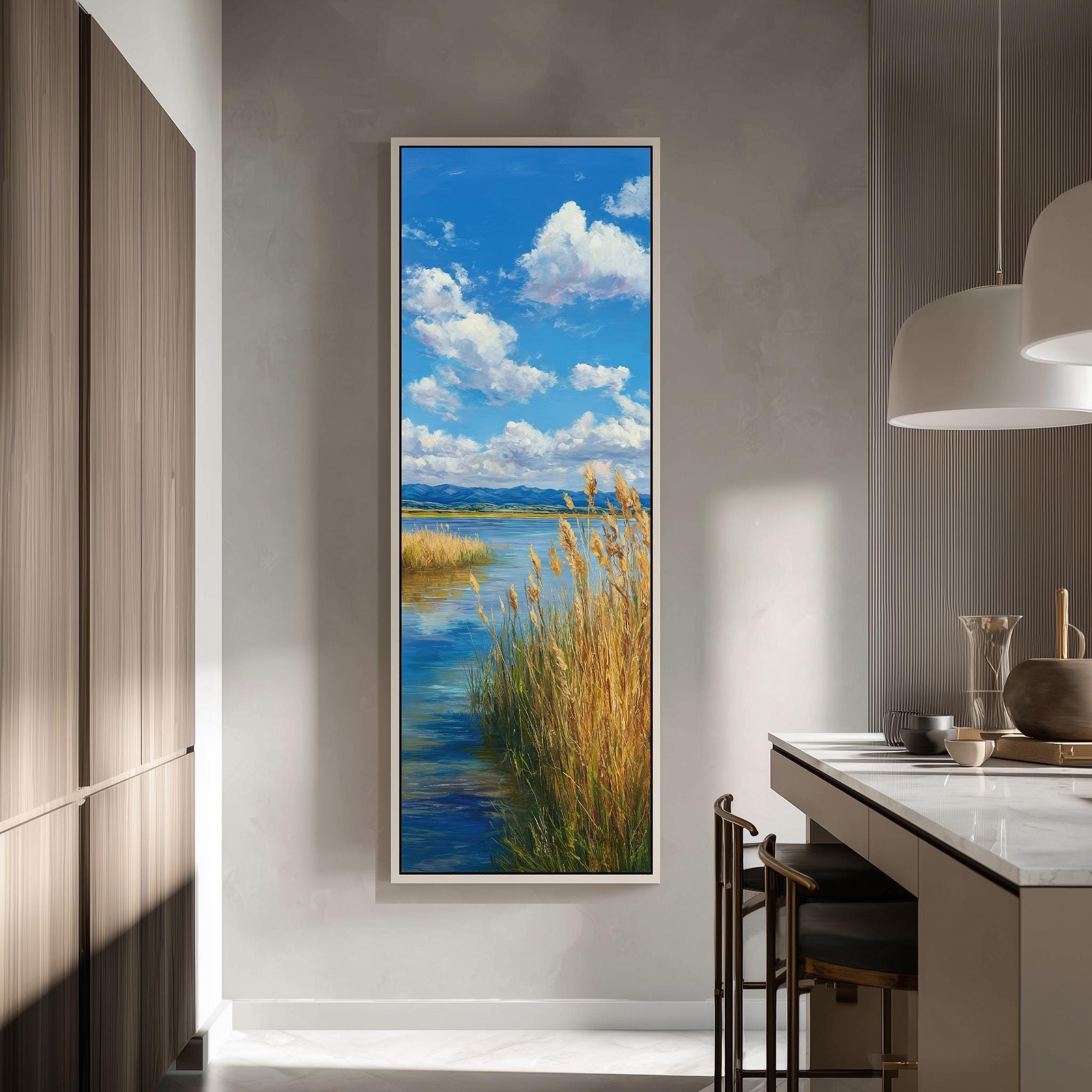 a large framed painting of a serene landscape with a river, grassy banks, and a blue sky with fluffy white clouds. The painting is hung on a wall in a modern kitchen with a countertop and chairs.