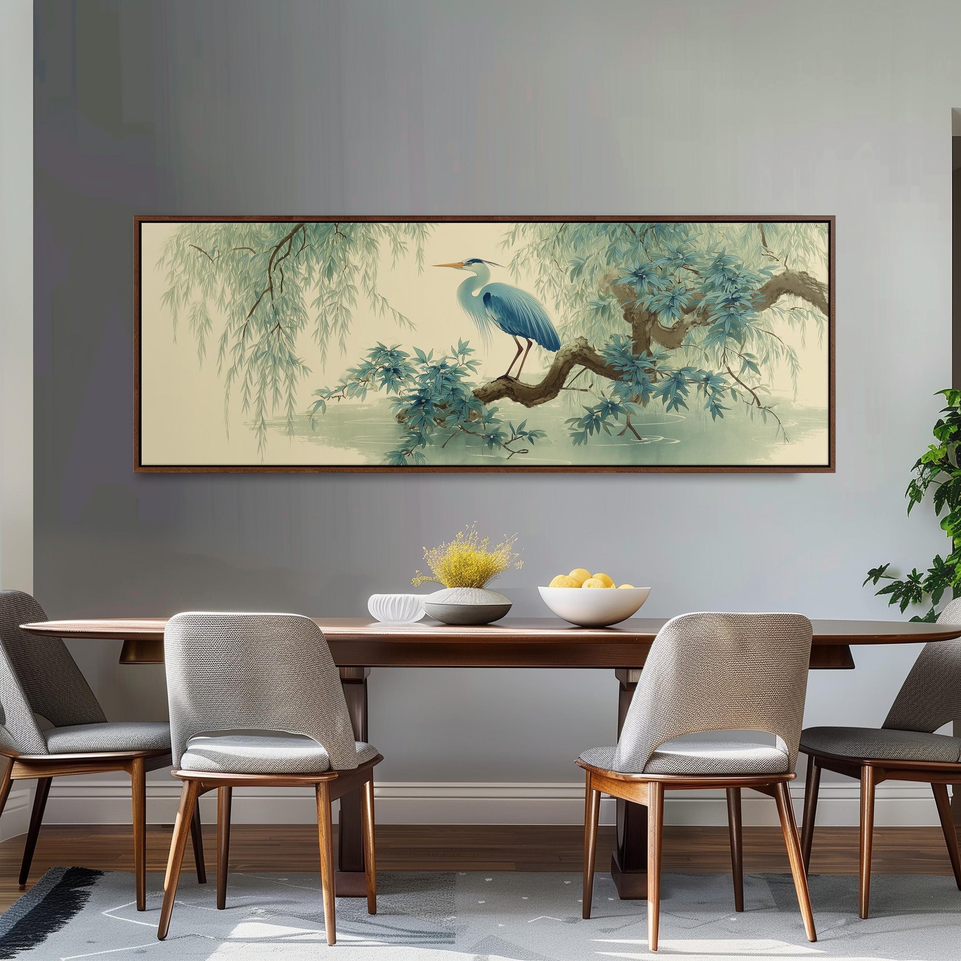 a dining room with a large painting of a blue heron perched on a branch above a wooden table. The table is set with a bowl of fruit and a vase of flowers, creating a cozy and inviting atmosphere. The room is decorated with gray chairs and a gray rug, contributing to the overall aesthetic.
