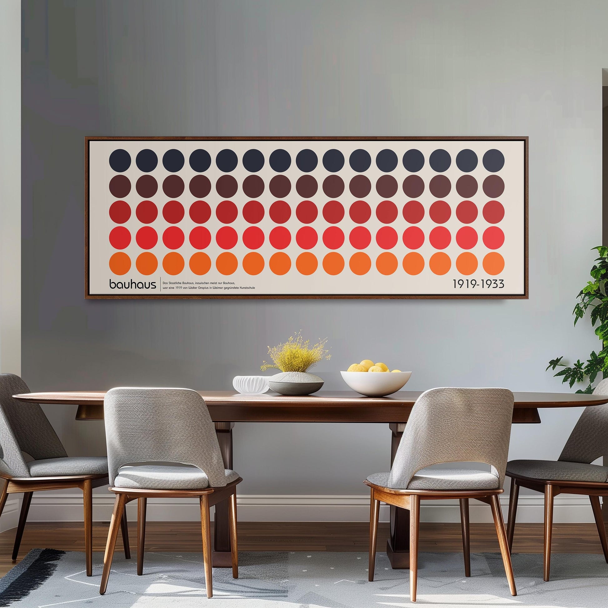 a dining room with a wooden table and four chairs. Above the table, there is a large framed poster with a repeating pattern of circles in various colors. The poster is hanging on a gray wall. On the table, there is a bowl of fruit and a vase of flowers.