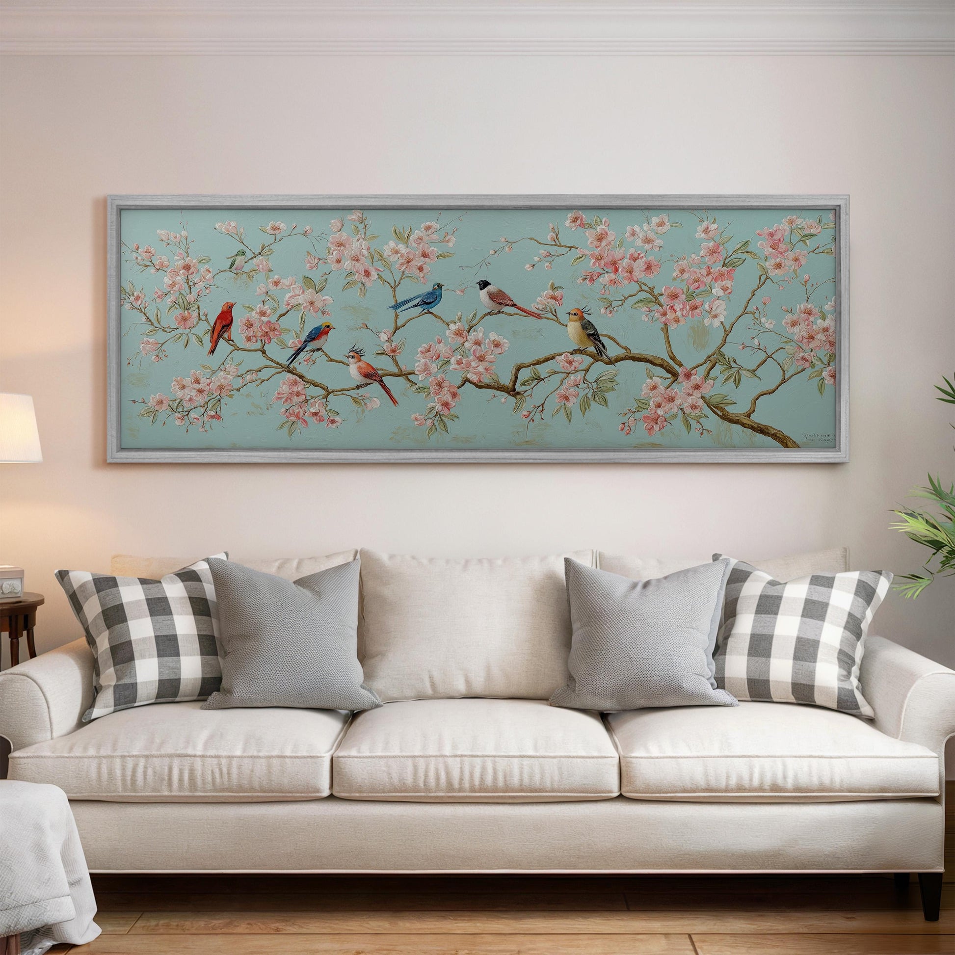 a large framed painting of birds perched on a branch with pink flowers, hanging on a wall above a white couch with gray and black plaid throw pillows.