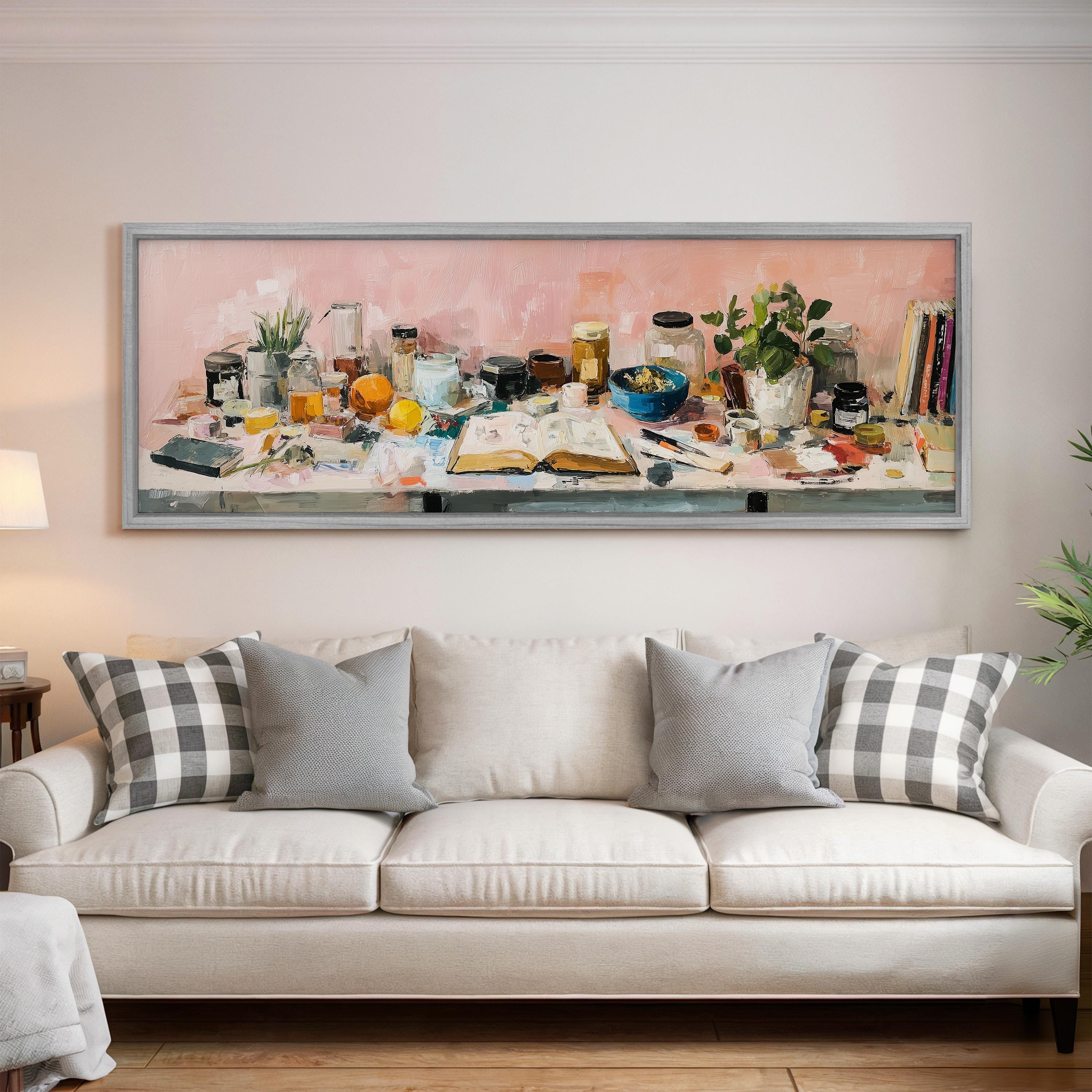 a large painting of a cluttered table with various objects, such as bottles, books, and a plant, hanging on a wall above a white couch with patterned pillows.