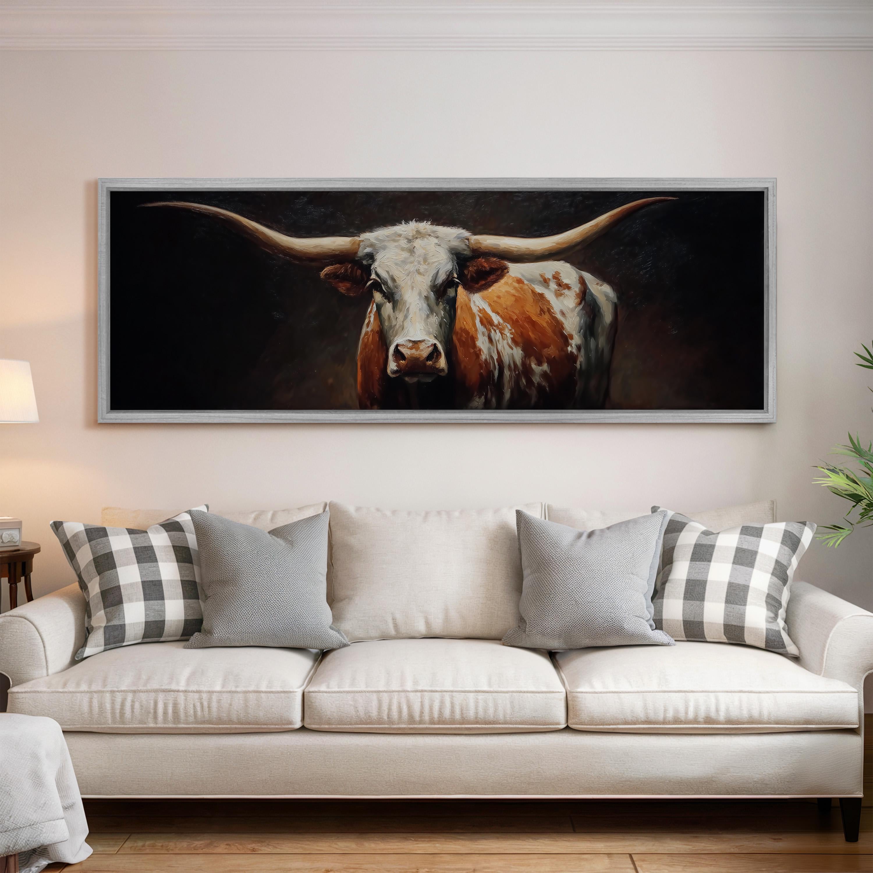 a large framed painting of a bull with long horns hanging on a wall above a white couch with plaid throw pillows.
