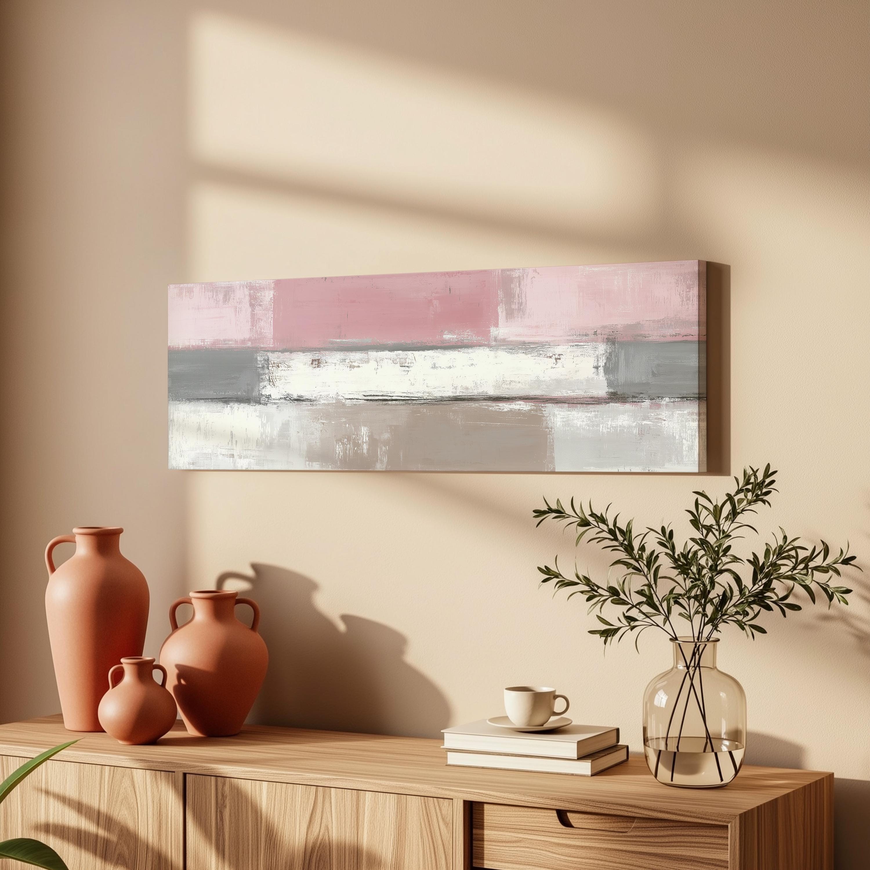 a wooden cabinet with a vase of greenery, a coffee cup, and a stack of books on top. Above the cabinet, there is a painting with a pink and gray color scheme.