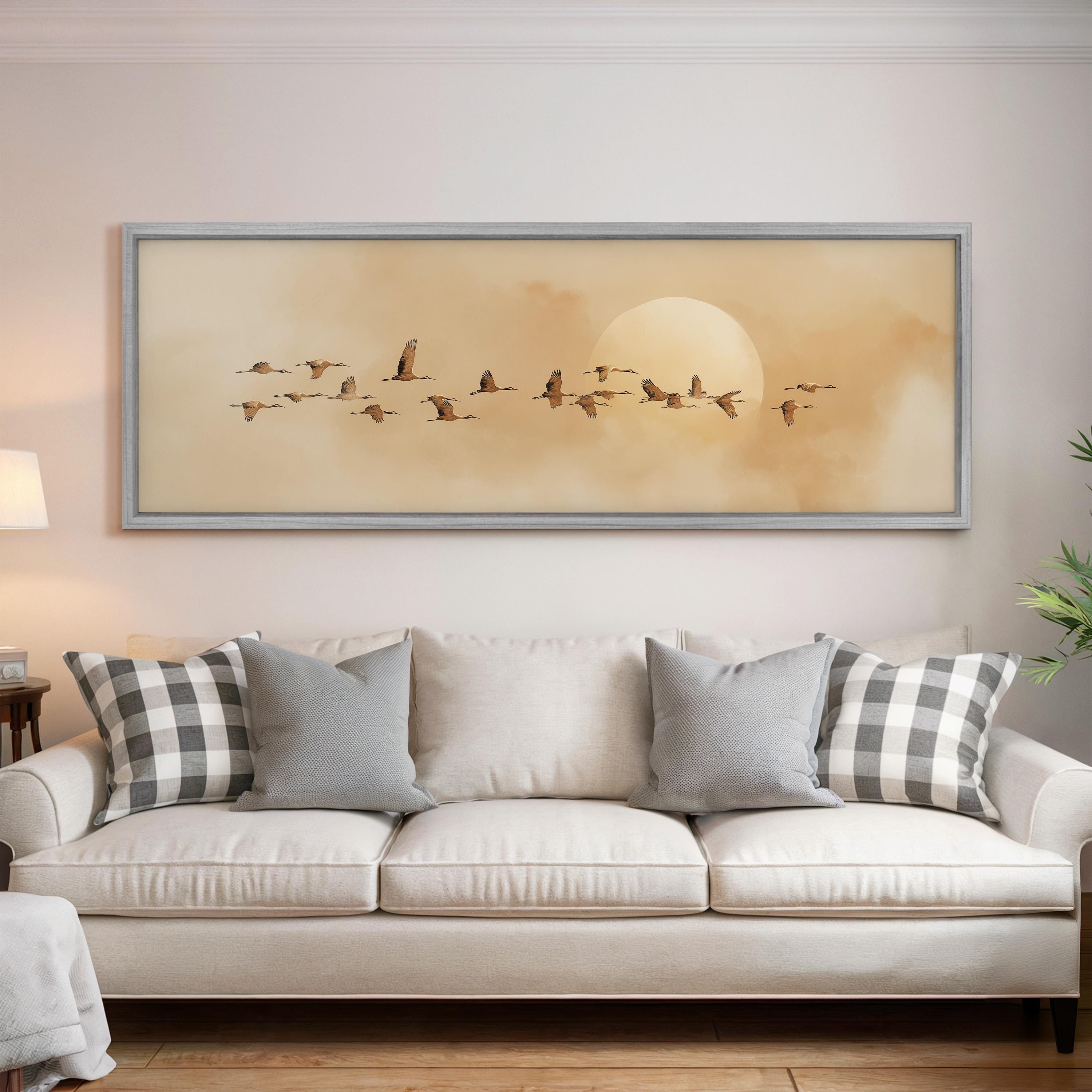 a large framed picture of a flock of birds flying over a sunset, hanging on a wall above a white couch with gray and white plaid throw pillows.