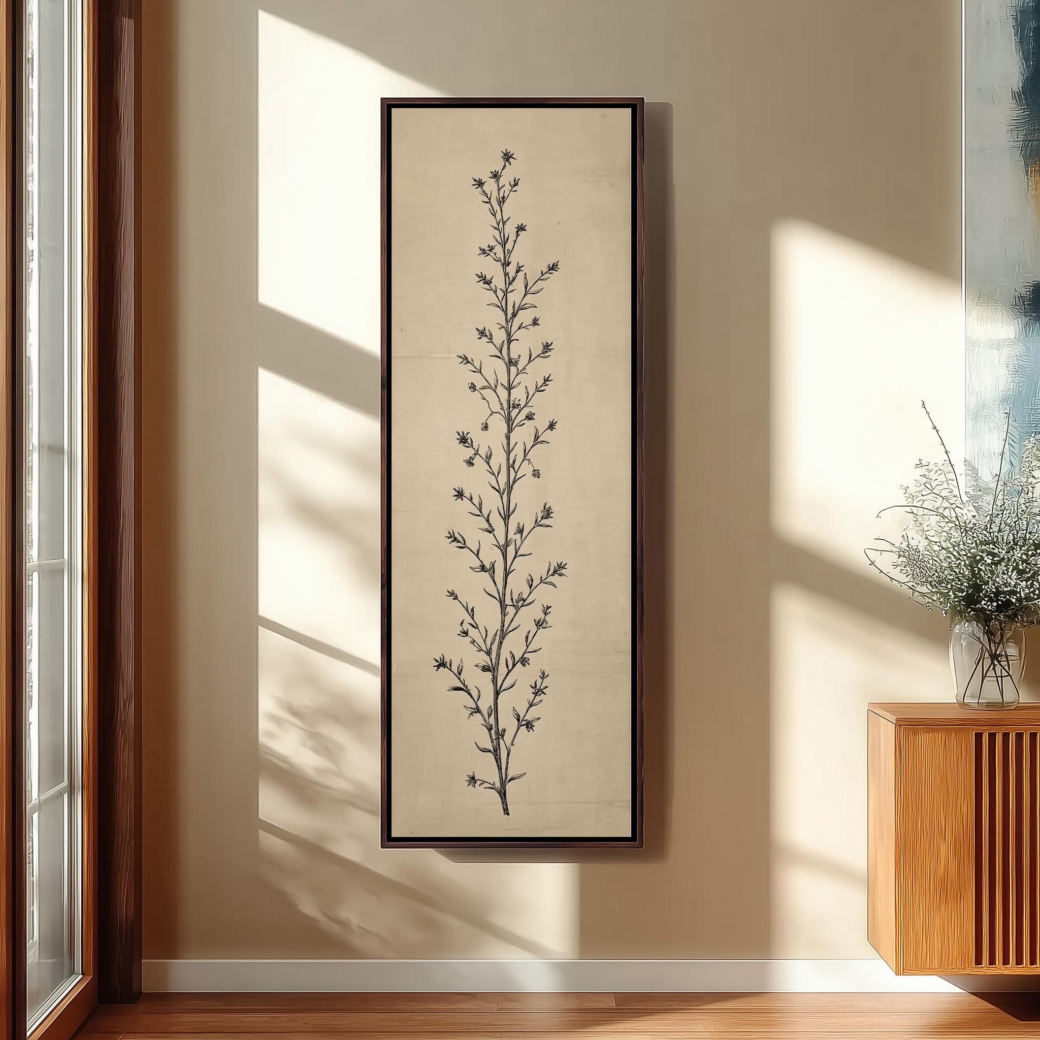a framed artwork of a tall plant with a vertical stem and numerous small leaves, hanging on a wall in a room with a wooden floor and a window.
