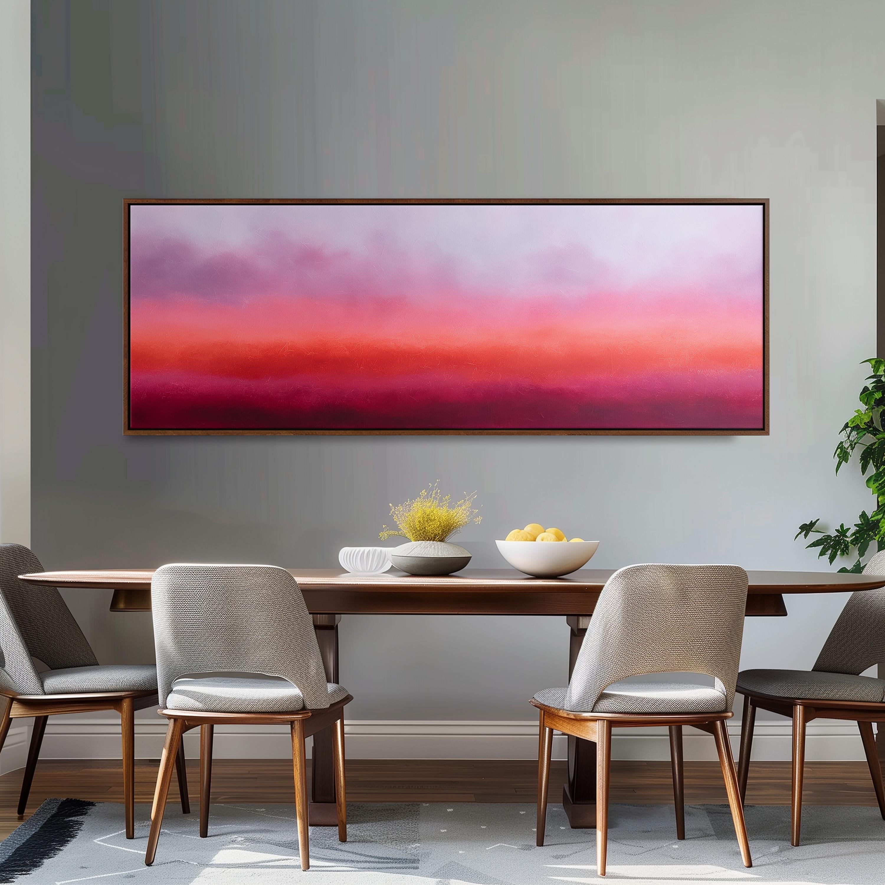 a dining room with a wooden table and four chairs. Above the table, there is a large painting depicting a sunset with a gradient of pink and orange colors. The painting is framed and hangs on the wall above the table.